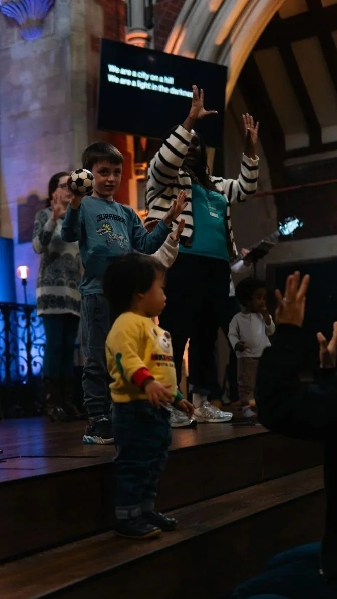 Children and adults raise their hands during an indoor event, with one boy holding a soccer ball beneath a projected message.