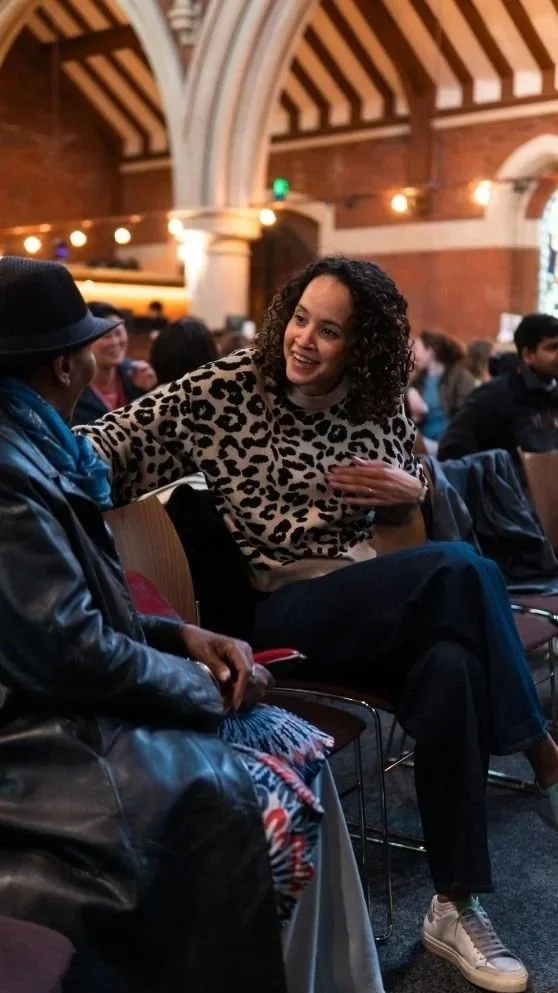 Woman in a leopard-print sweater chatting with a person in a black hat at a crowded indoor event.