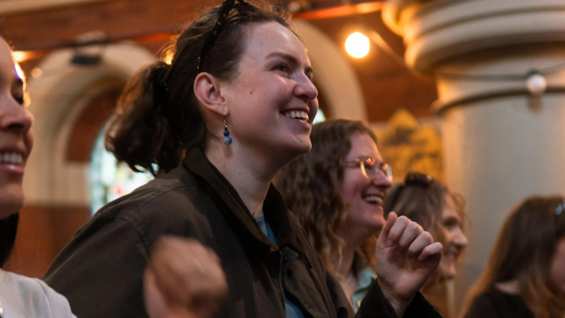 Smiling woman with blue earrings among an enthusiastic crowd at an indoor event