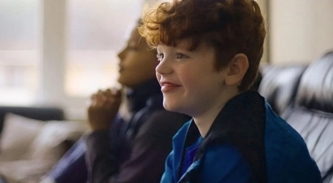 Smiling red-haired boy sitting on a couch with an adult in the background