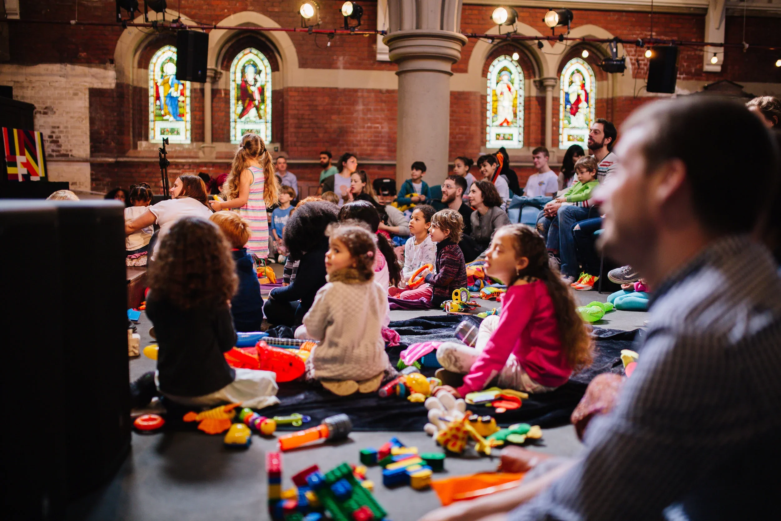 Children and families gather for a colorful musical activity in a church with stained-glass windows.
