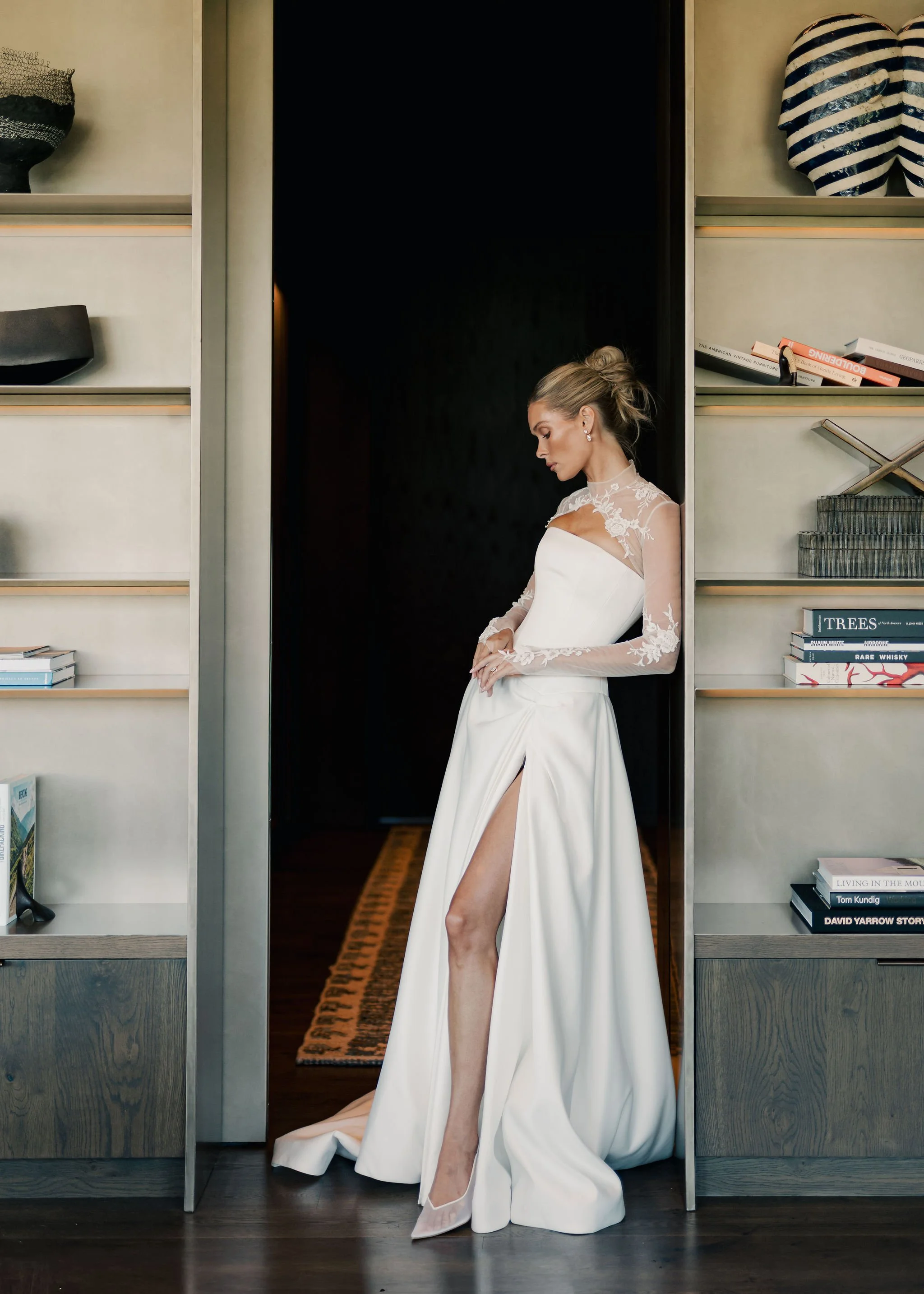Woman in a white wedding dress standing in between two bookshelves, her hands clasped, viewing downward with a side profile in an elegant room.