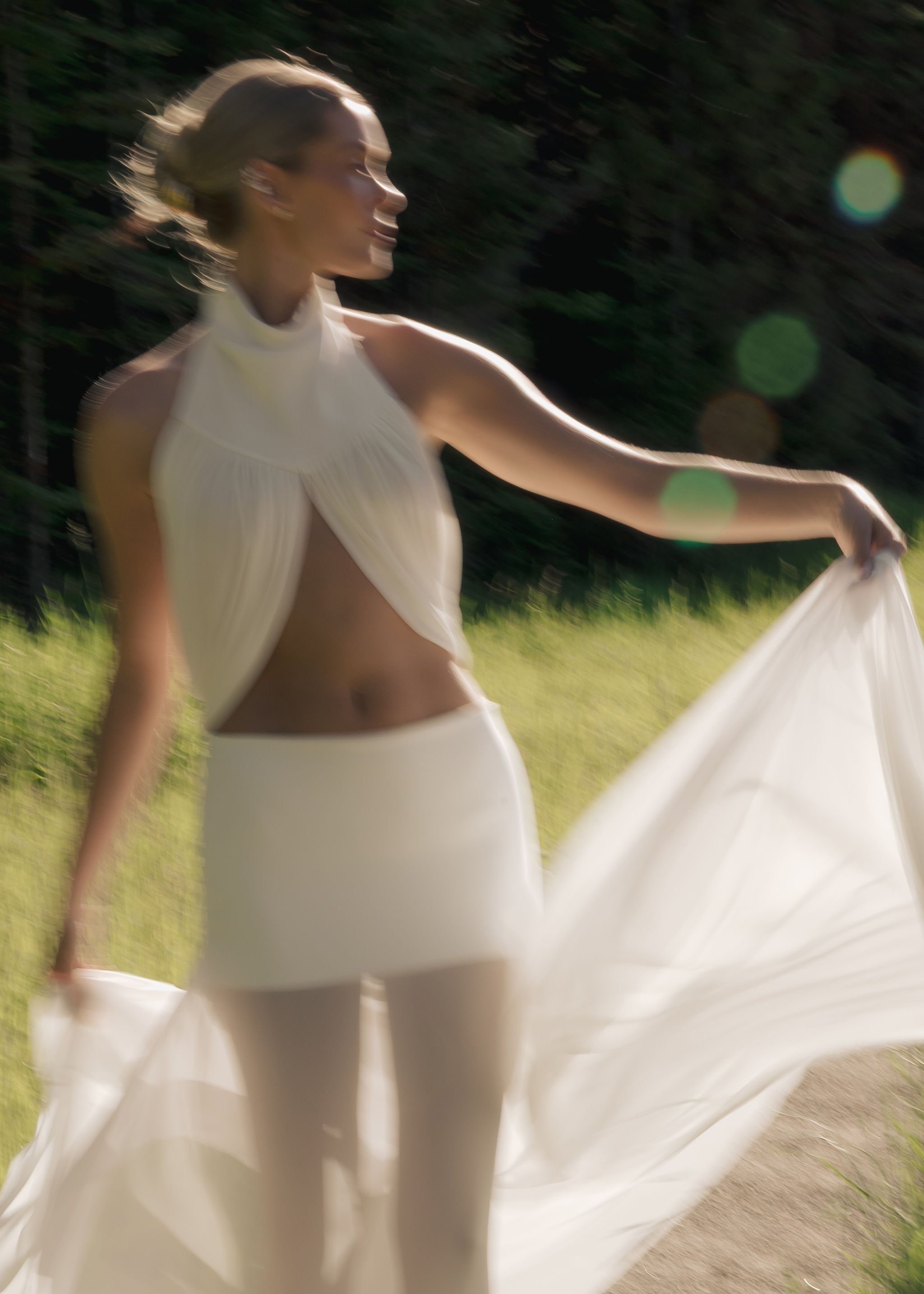 A woman with blonde hair in a bun outdoors, wearing a white sleeveless top and a flowing white skirt, with sunlight and lens flare effects.