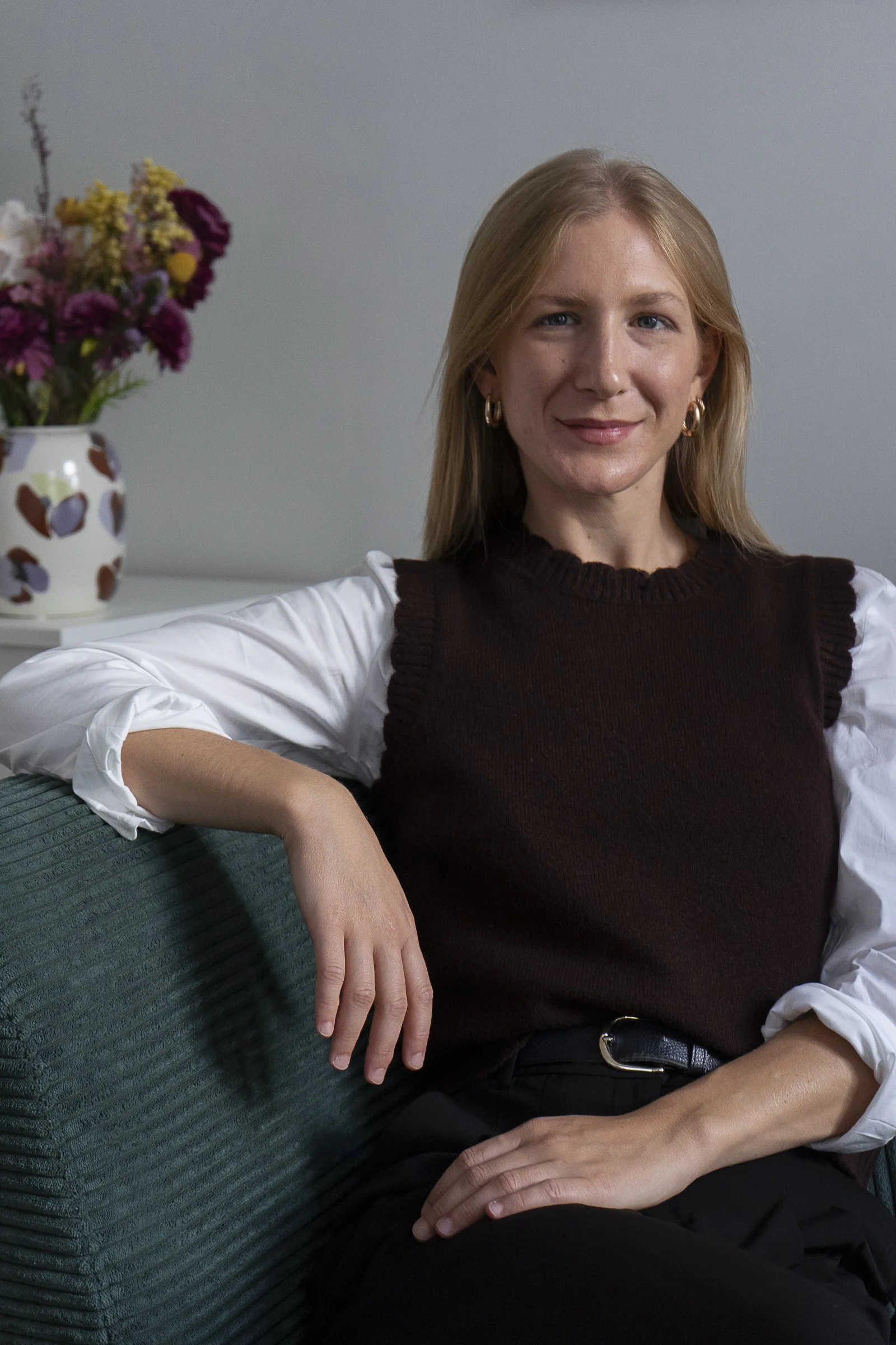 Eine Frau sitzt entspannt auf einem grünen Sofa, trägt eine weiße Bluse und eine dunkelbraune Weste, hat blonde Haare und trägt Goldohrringe, im Hintergrund eine Vase mit bunten Blumen.