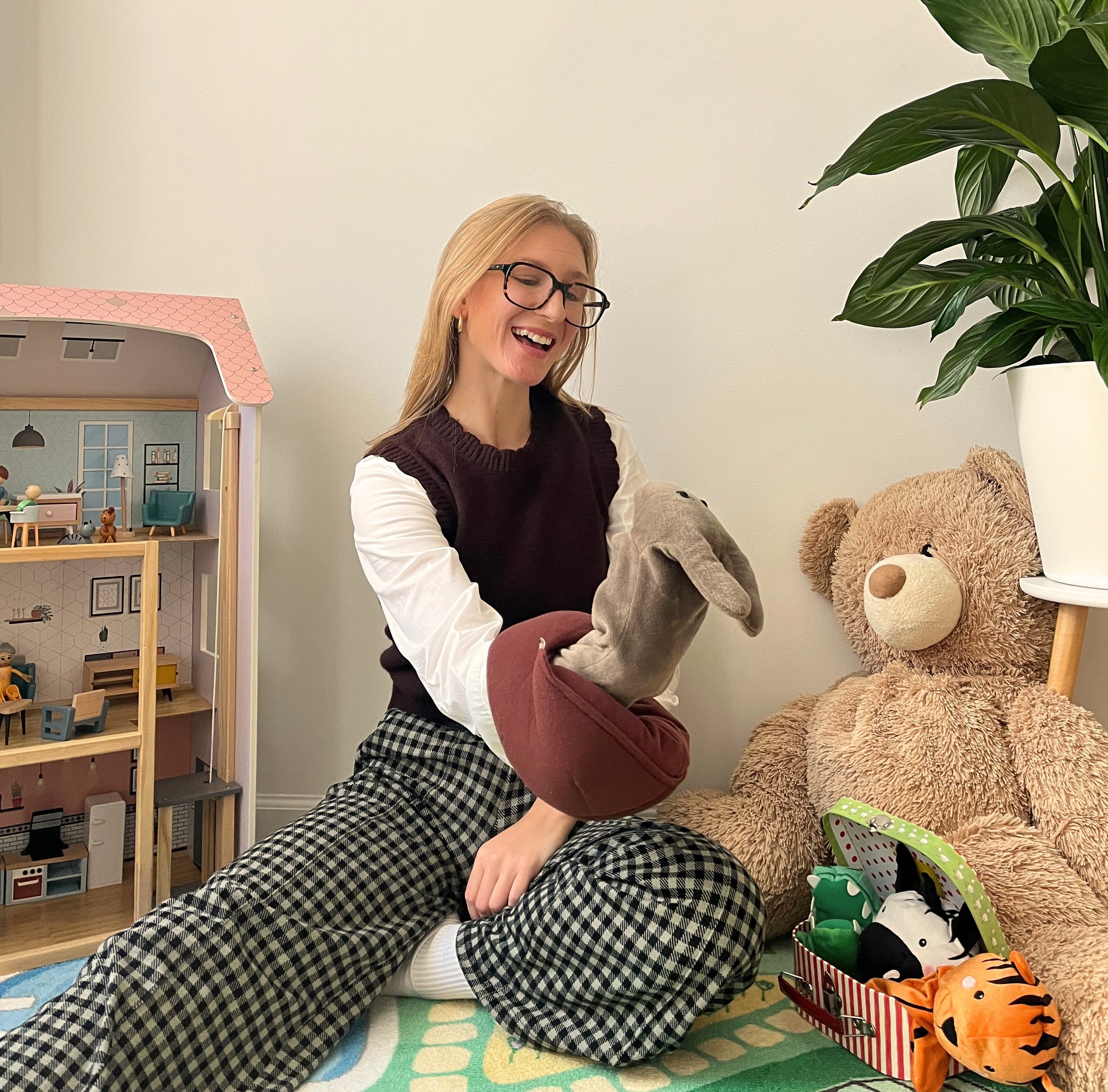 Lachende Frau mit Brille hält ein Stofftier in einem gemütlichen Kinderzimmer neben einem großen Teddybären.