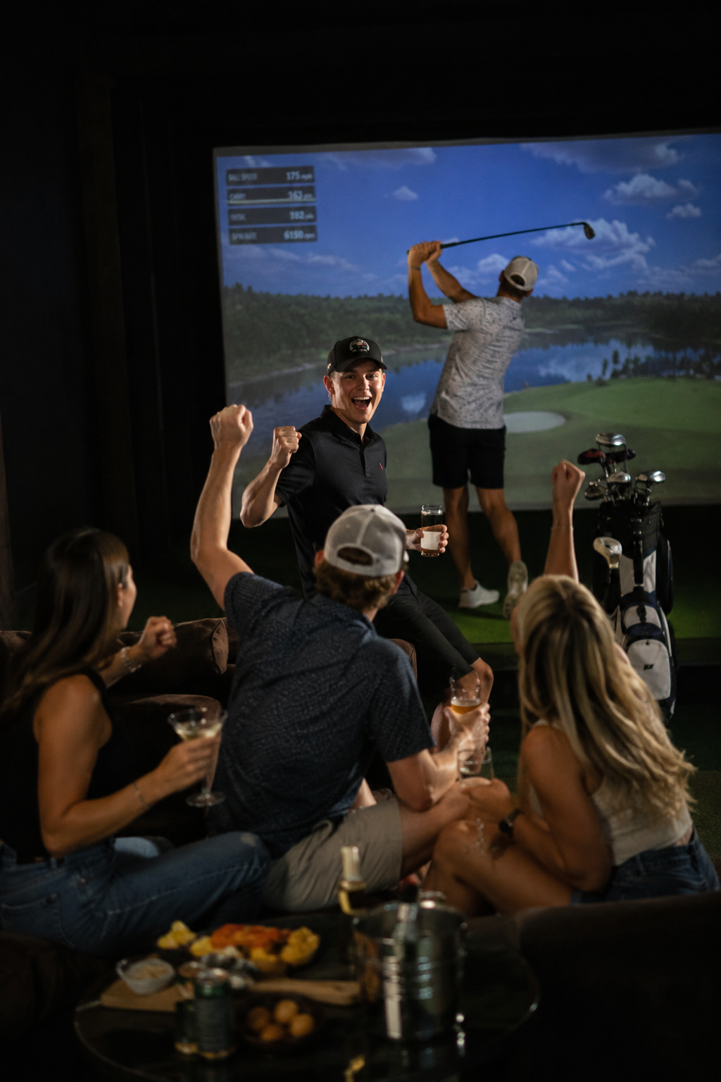 Friends watching a golf game on a screen at a bar, celebrating, holding drinks, with snacks on a table in the foreground.