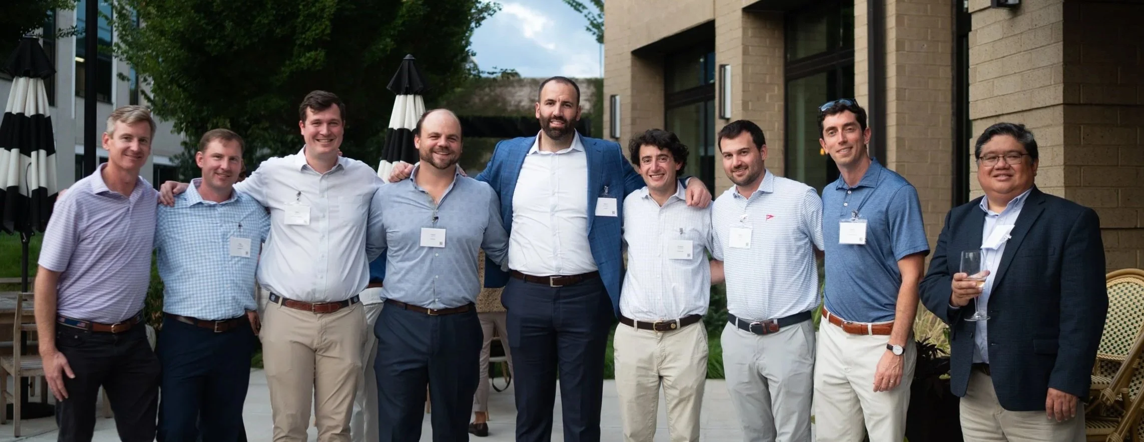 Group of nine men standing outdoors at a business event, smiling, with some wearing name tags, and one holding a glass of wine.