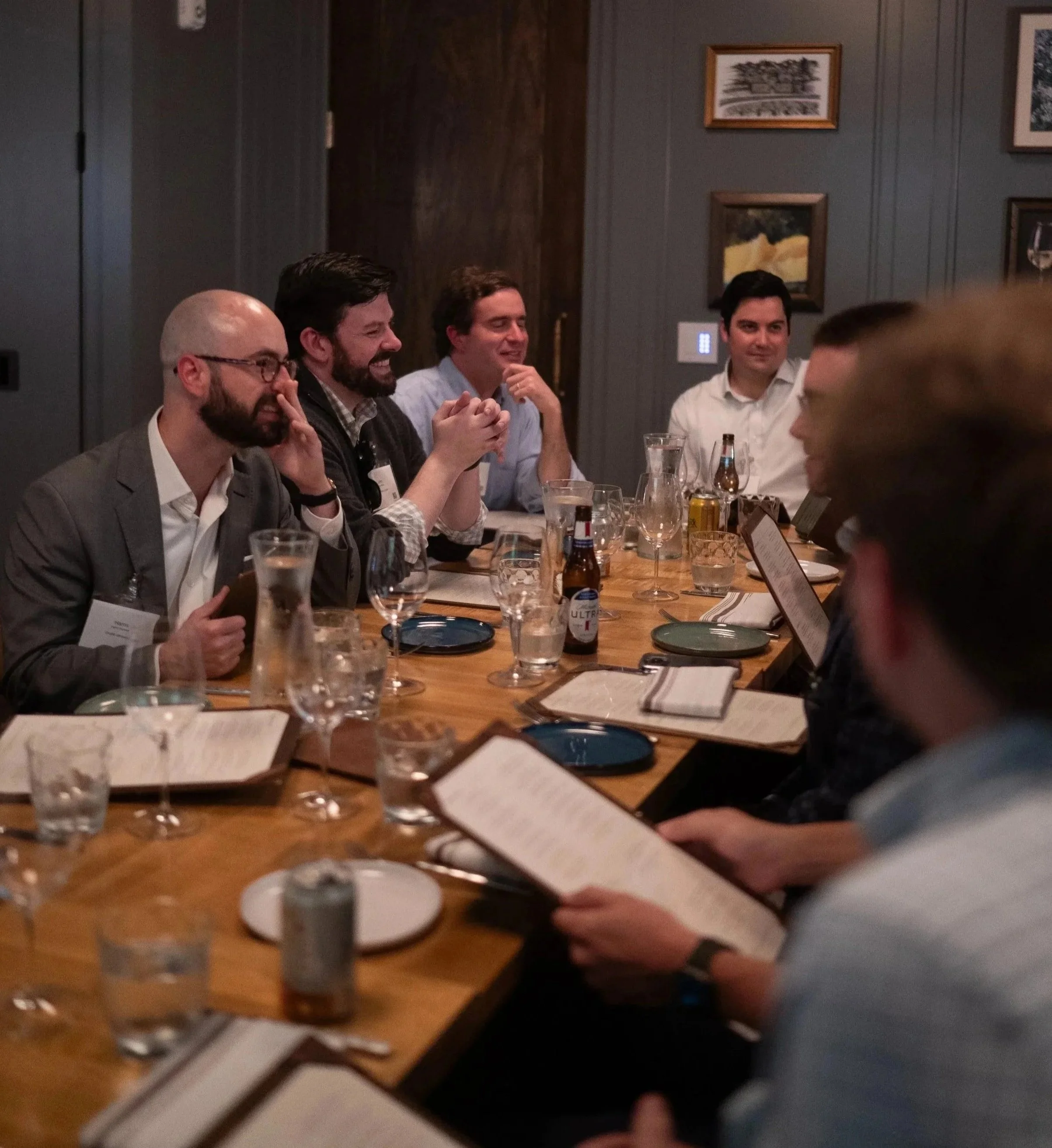 People sitting at a long table during a social gathering or dinner, with drinks and menus in front of them, in a dimly lit room with framed artwork on the walls.