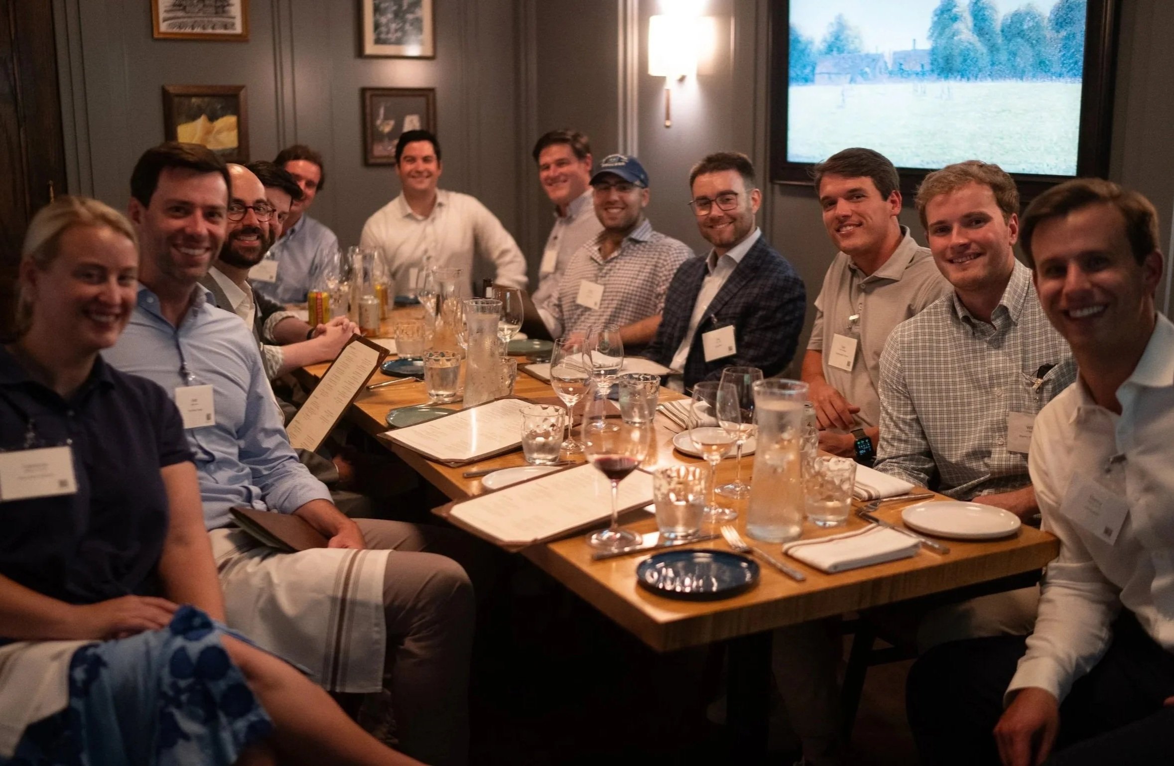 A group of people sitting around a rectangular wooden dining table in a dimly lit restaurant, smiling, with wine glasses, menus, and drinks on the table, and a large flat-screen TV showing an outdoor scene on the wall in the background.