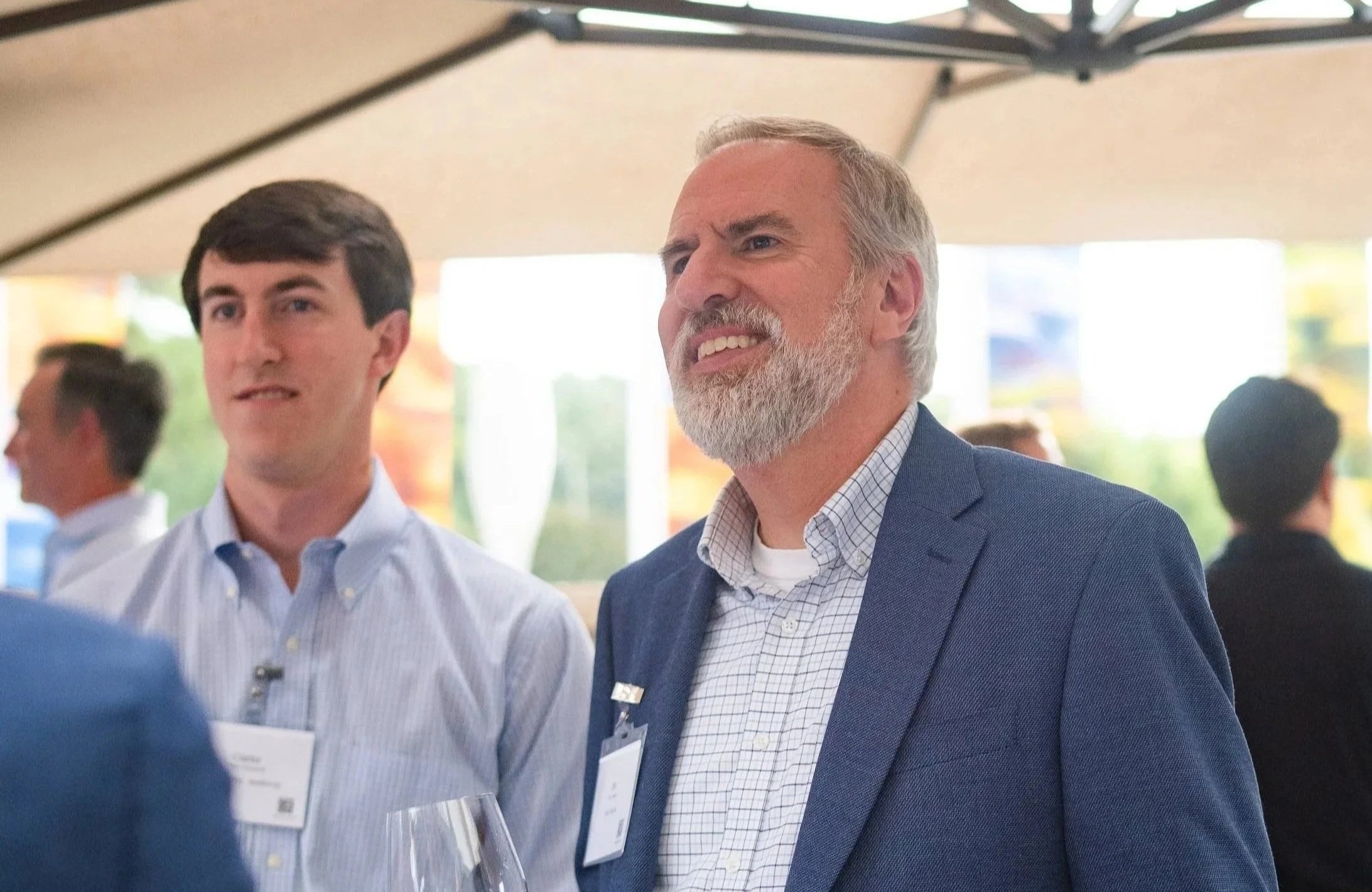 Two men in formal business attire standing at an indoor event, engaging in conversation. The man in the foreground has white hair and a beard, wearing a blue blazer, while the other man has dark hair, wearing a light blue shirt. There are blurred people and displays in the background.