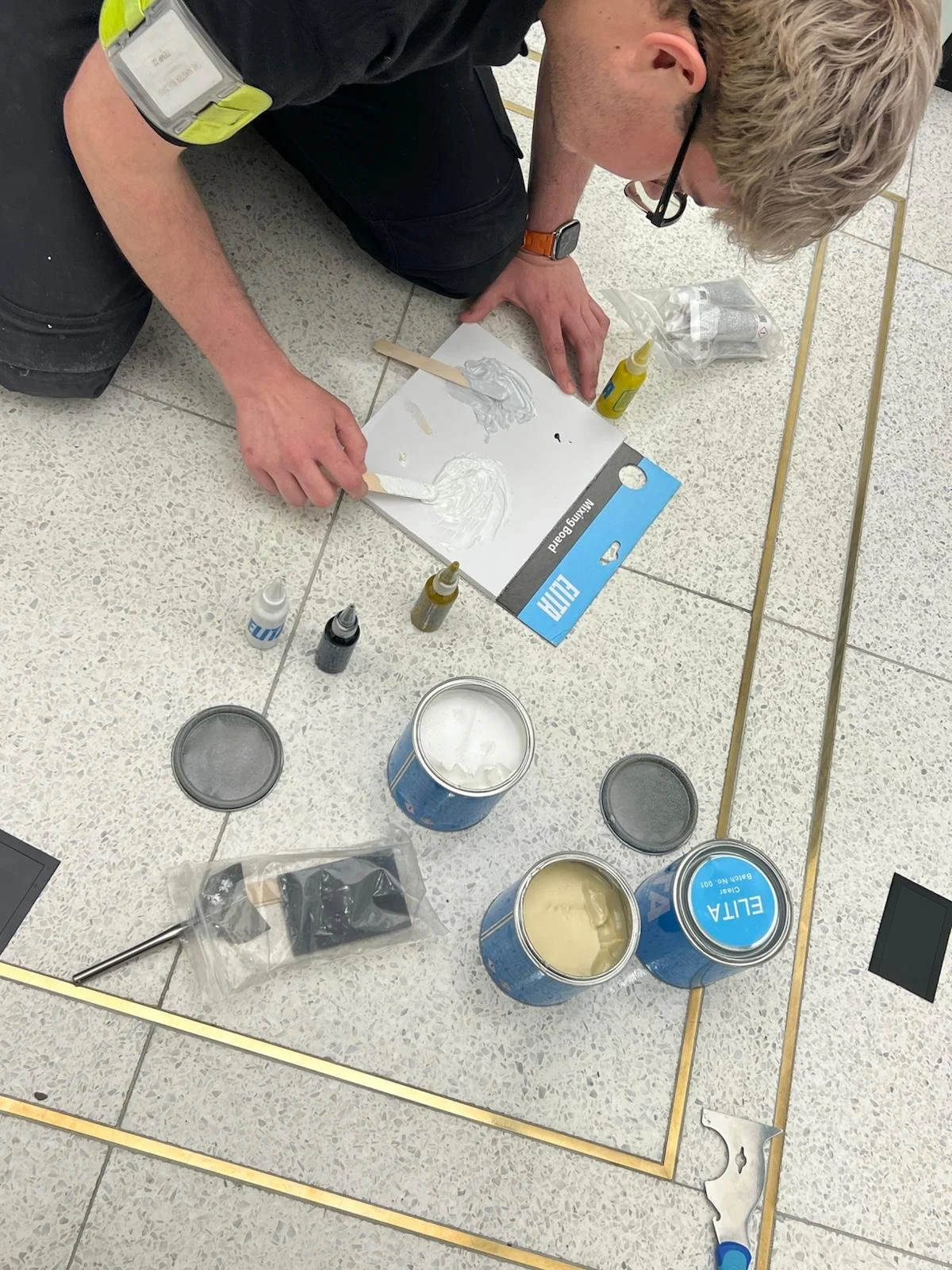 A person kneeling on terrazzo floor creating artwork with white modeling clay or plaster, surrounded by open cans of paint or plaster, bottles of glue or liquid, and various tools.