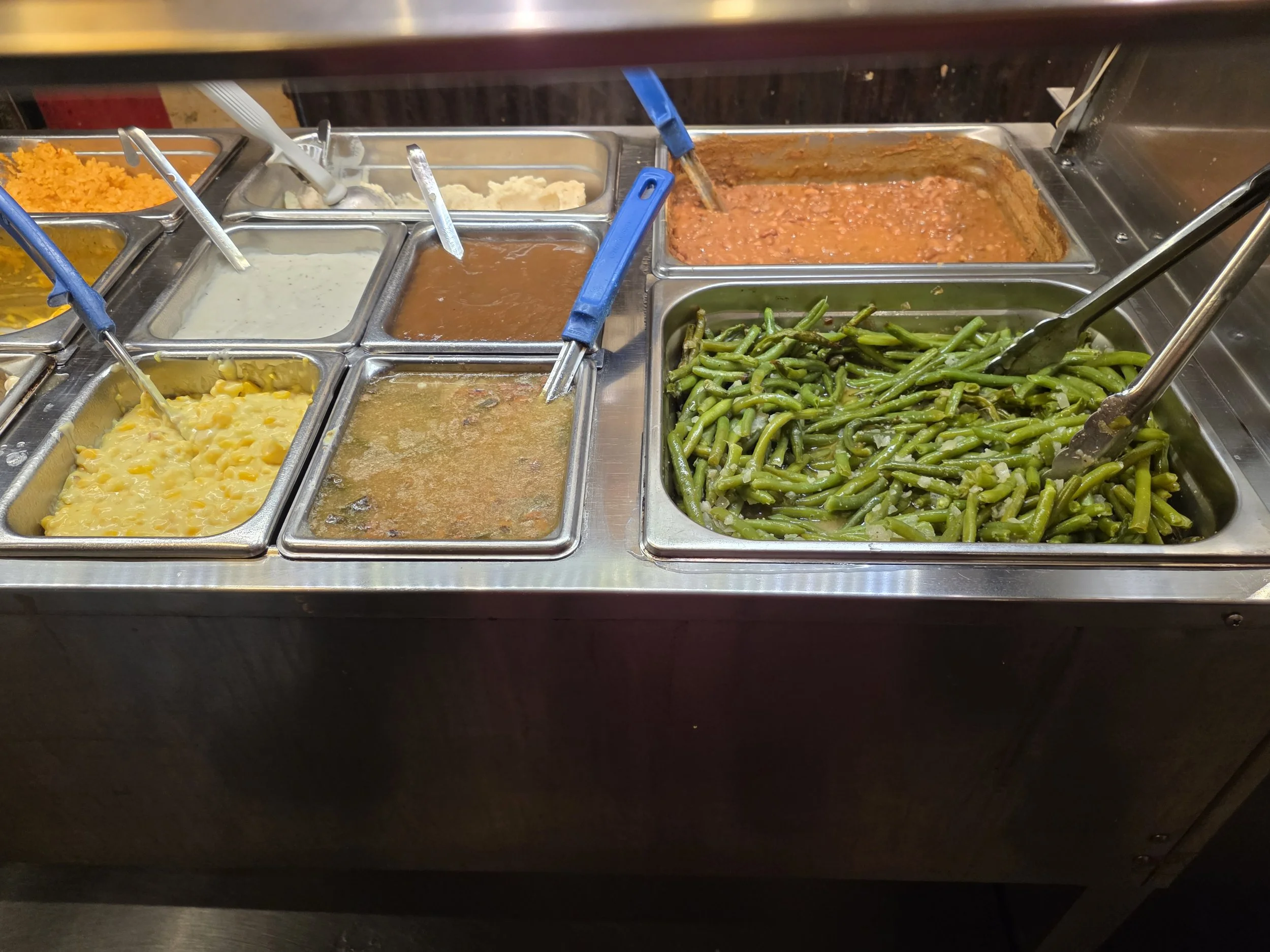 A cafeteria buffet with various food trays including green beans, mac n cheese, and baked beans.