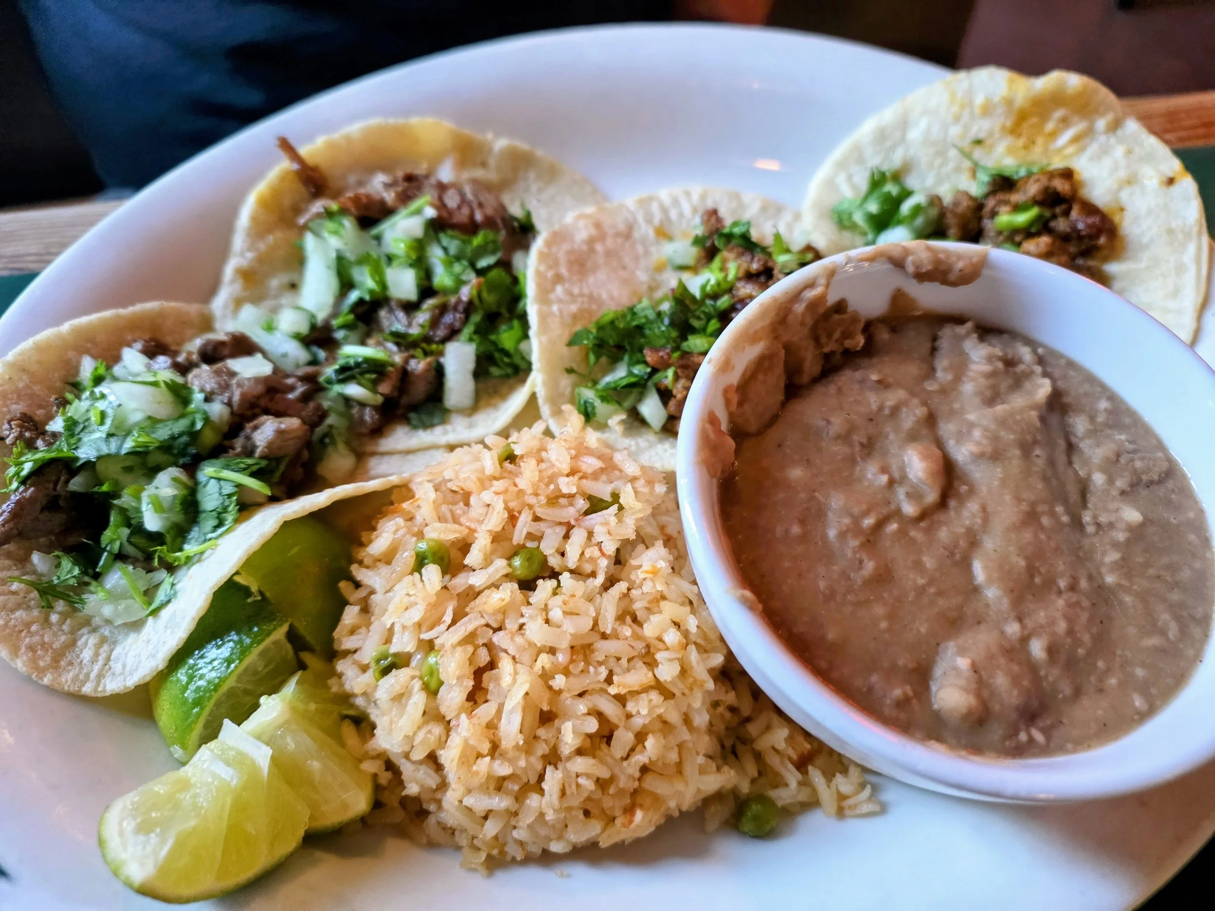 Close-up of Mexican street tacos with minced meat, chopped onions, cilantro, and lime wedges on a white plate, along with rice and a side of beans.