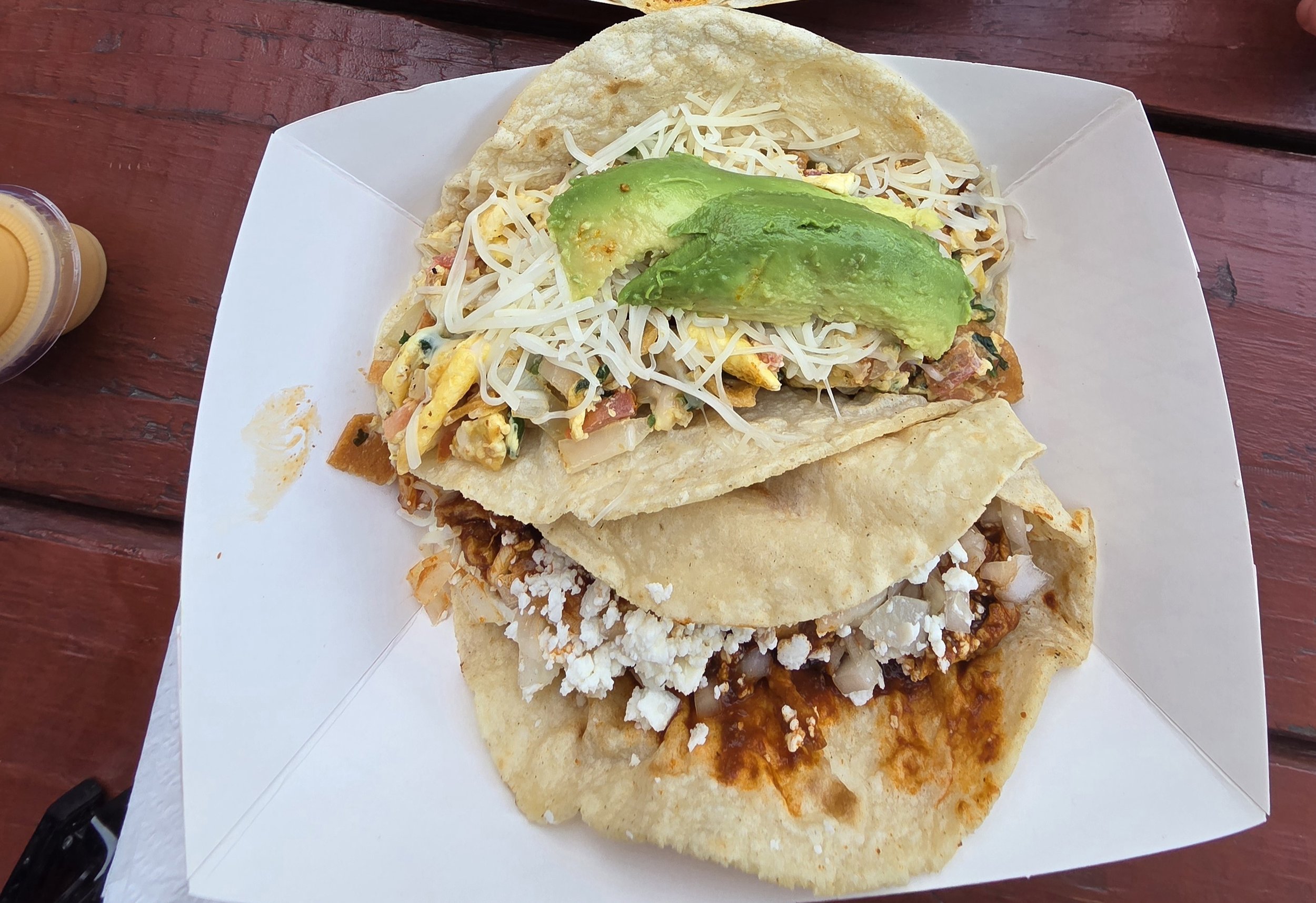 Two tacos on a white paper tray with avocado slices on top and shredded cheese with eggs and tortillas called migas, a chicken mole taco in a paper boat, served on a wooden table.