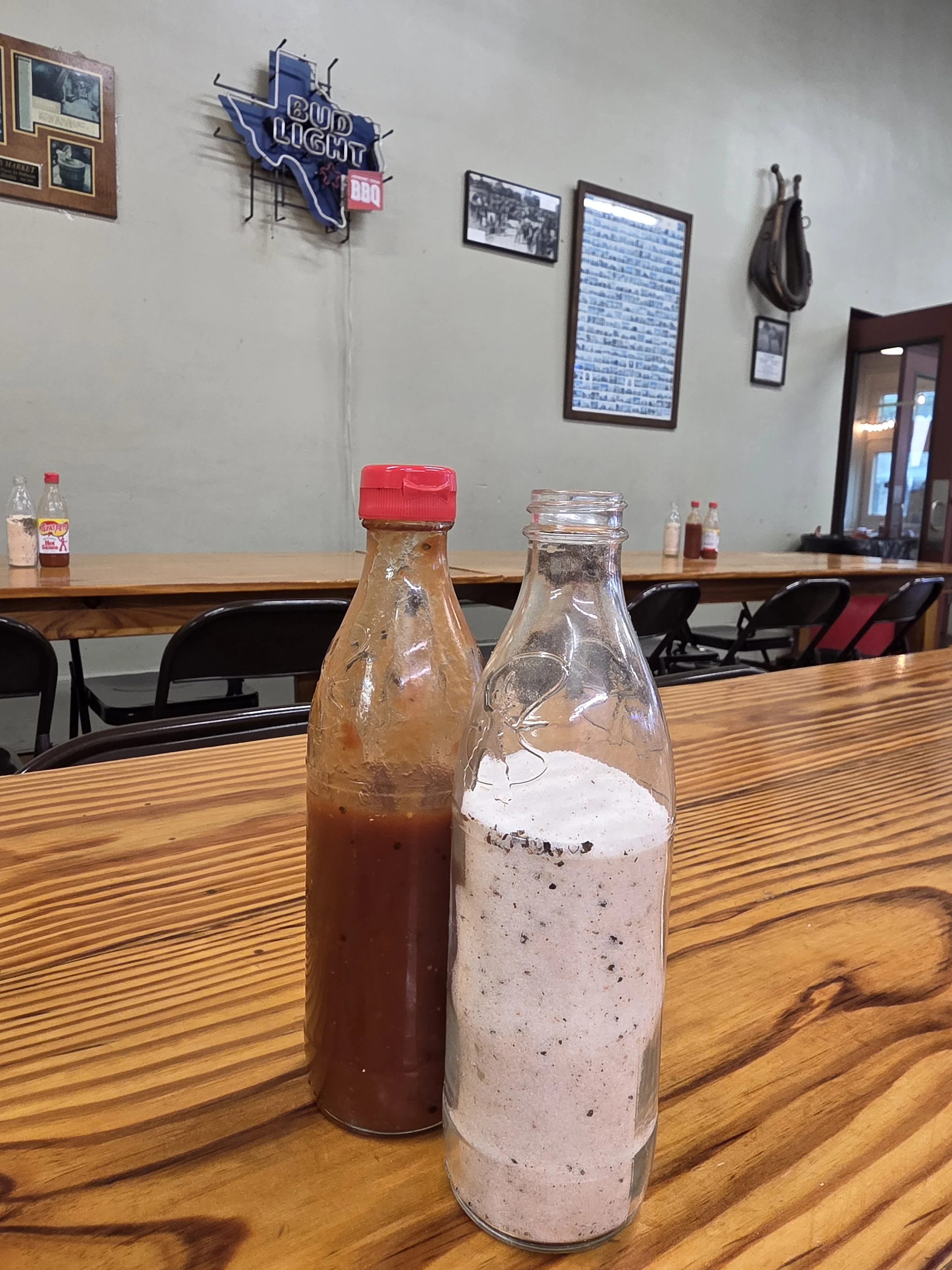 Two glass condiment bottles, one with a red cap containing hot sauce and the other with a clear cap containing a bbq seasoning salt, on a wooden table in a restaurant with framed pictures and beer signs on the wall.