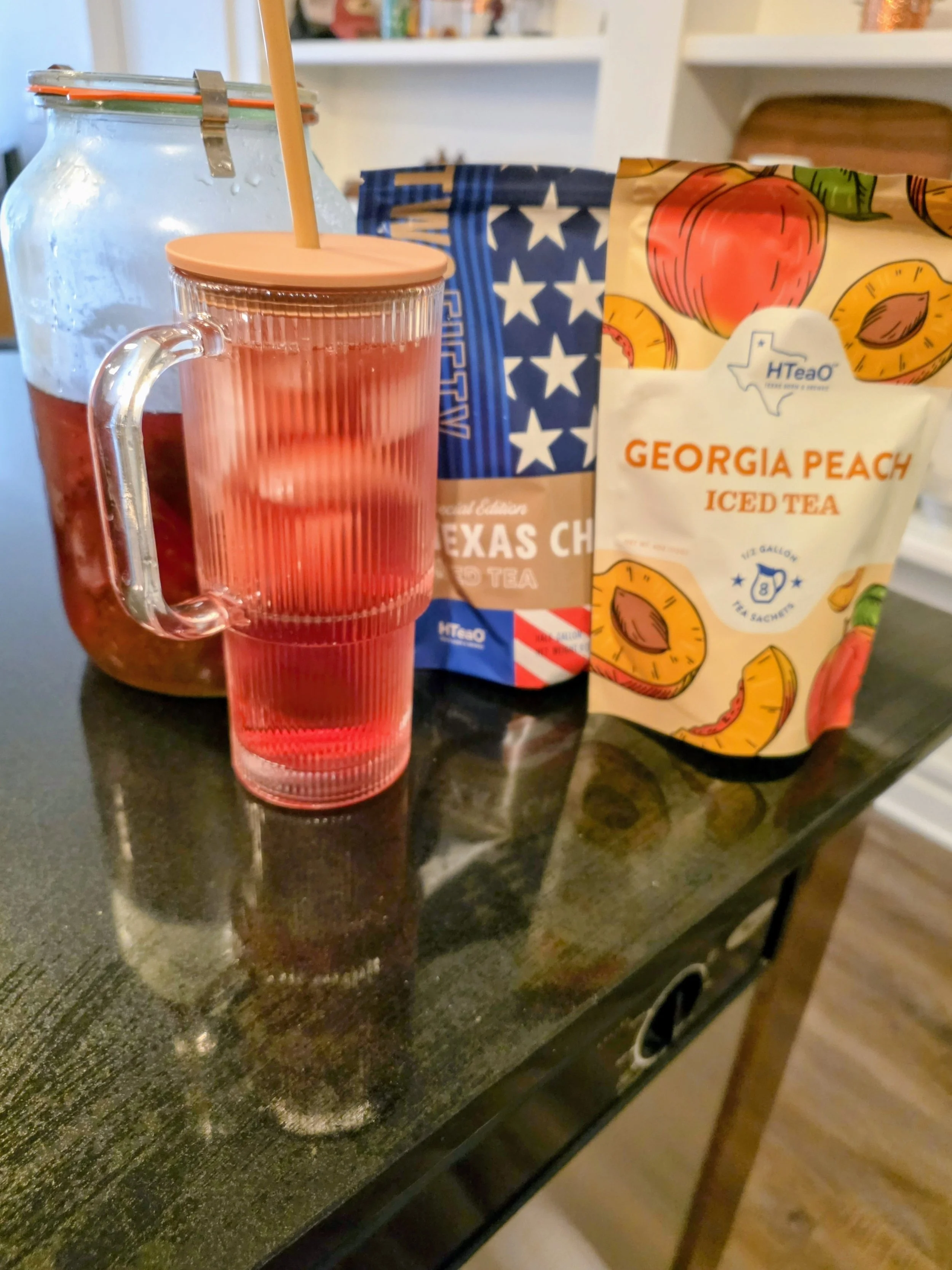 A glass pitcher with a pink lid and pink straw, a bag of Georgia Peach iced tea, and a bag of Texas chili tea on a black countertop.
