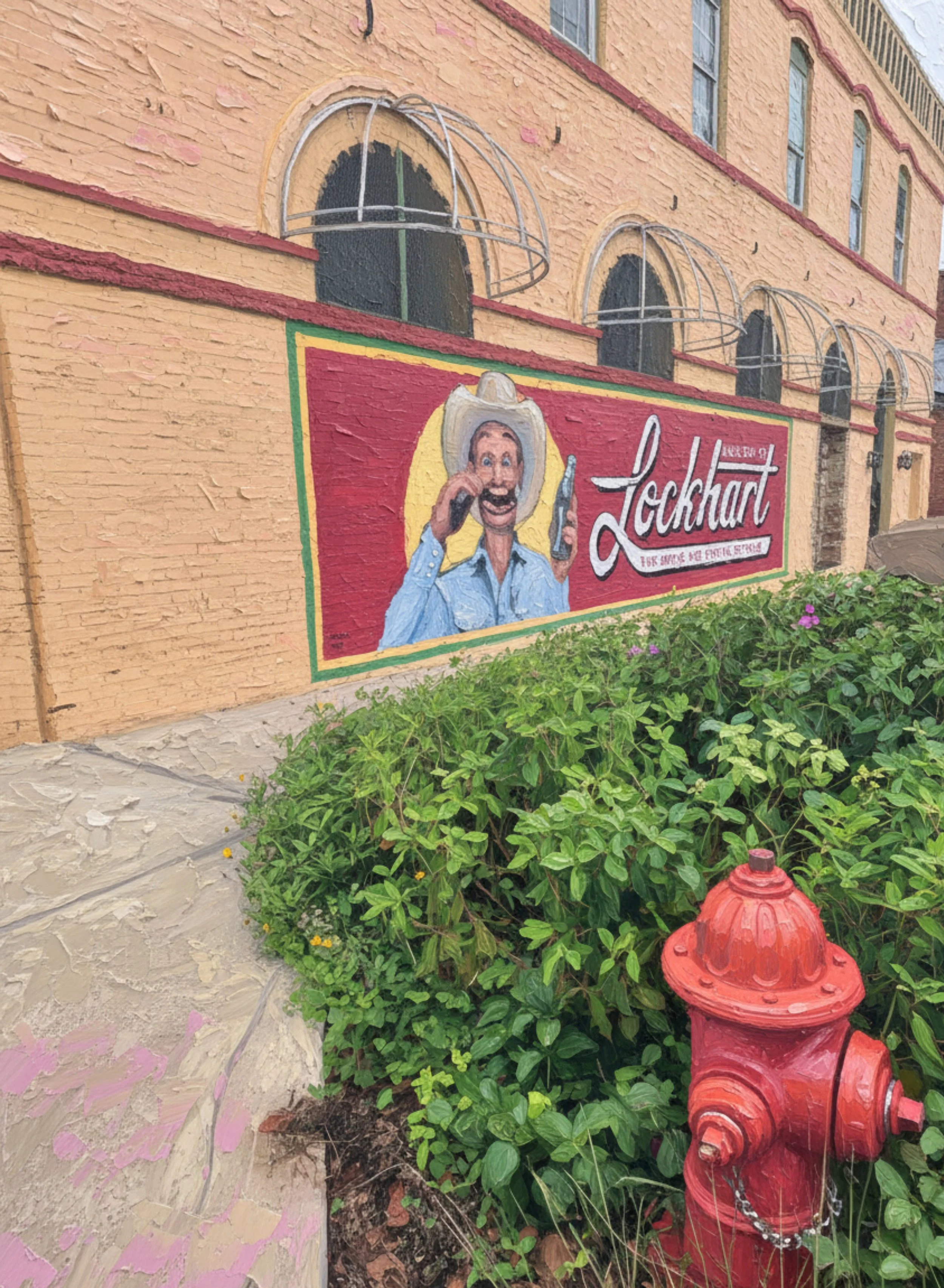 A mural of a man in a cowboy hat , painted on a yellow brick wall of a building. In front of the wall, there is a red fire hydrant surrounded by green bushes, and pink flowers are visible near the bottom of the image.