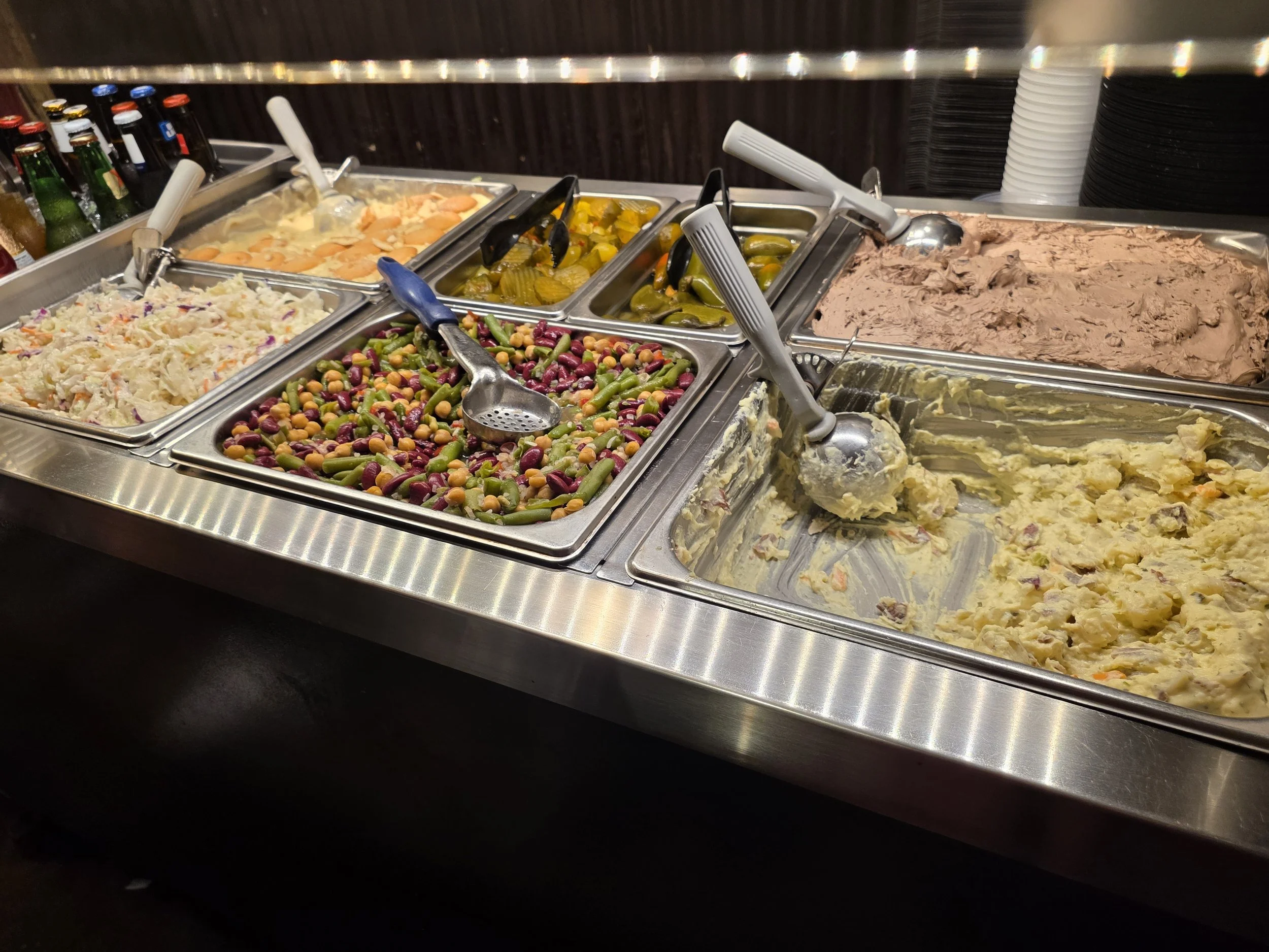 Cold cafeteria style options with various dishes including potato salad, mixed vegetables, pickles, pudding and other cold side dishes, along with bottled beverages and stacked plates in the background.