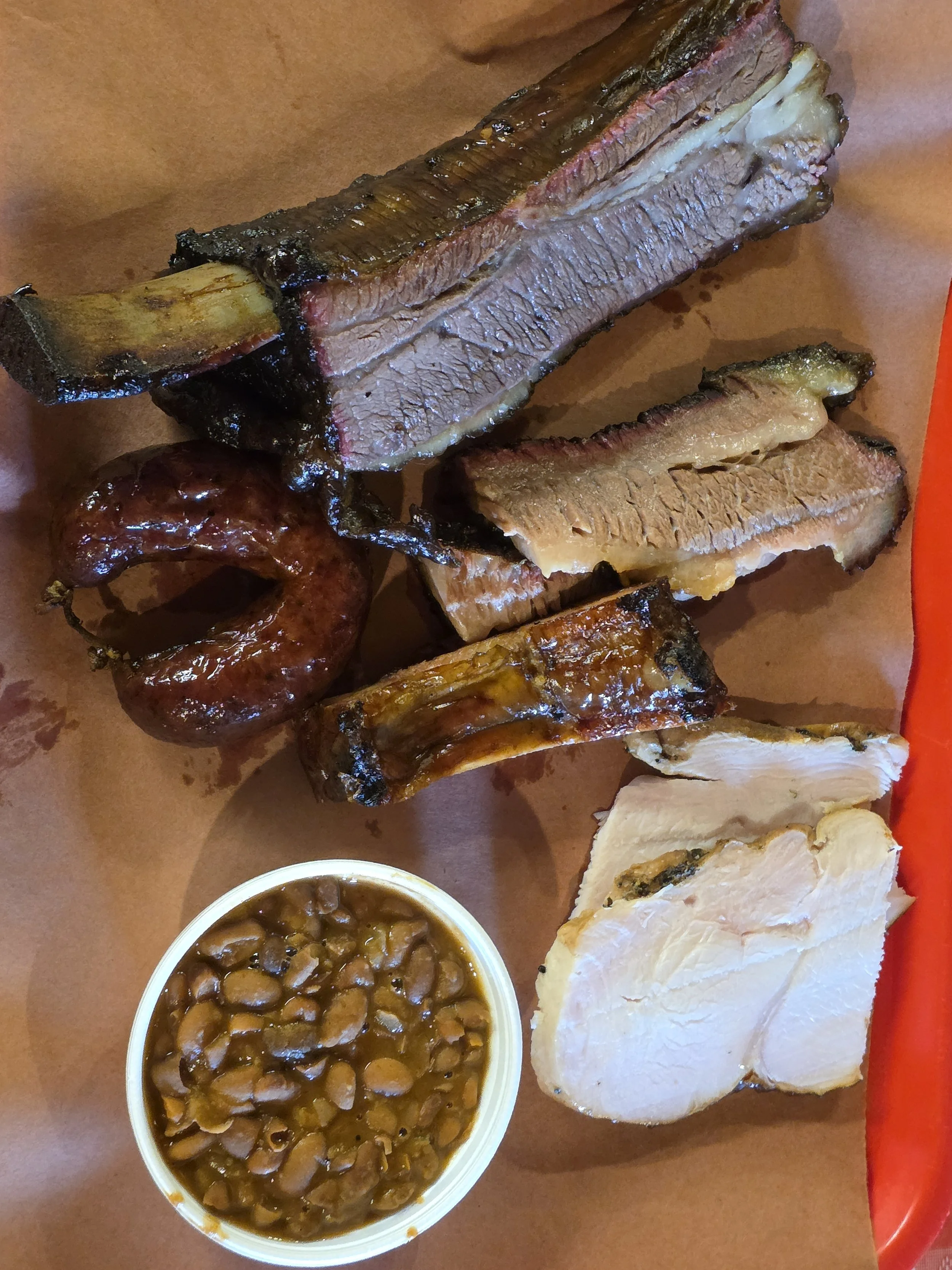 Barbecue ribs with beans, sliced chicken turkey, sausage, brisket and pork rib on a brown paper meal tray.