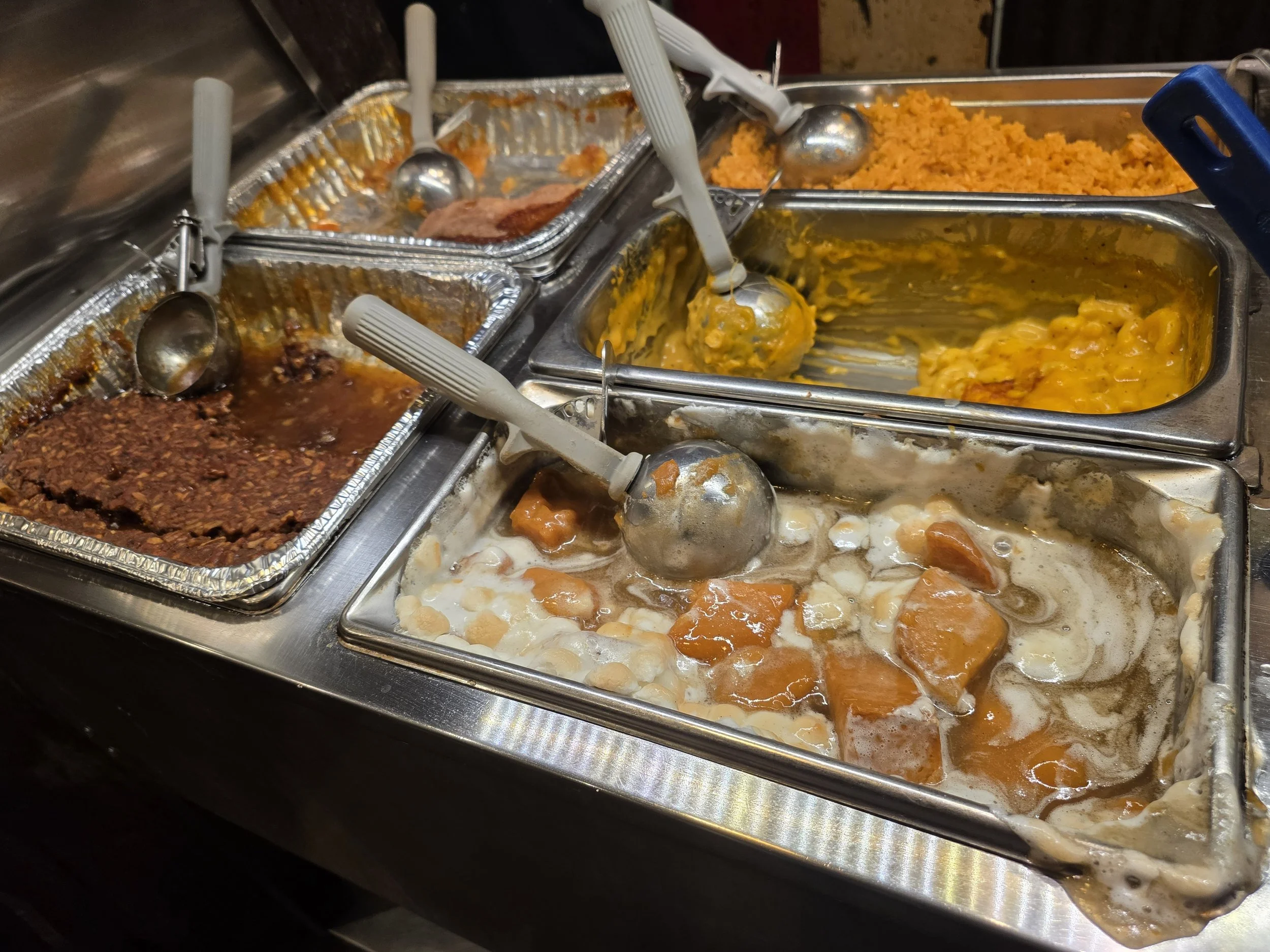 Various trays of hot sides, including sweet potatoes, vegetables, and baked dishes, with serving utensils in metal containers.