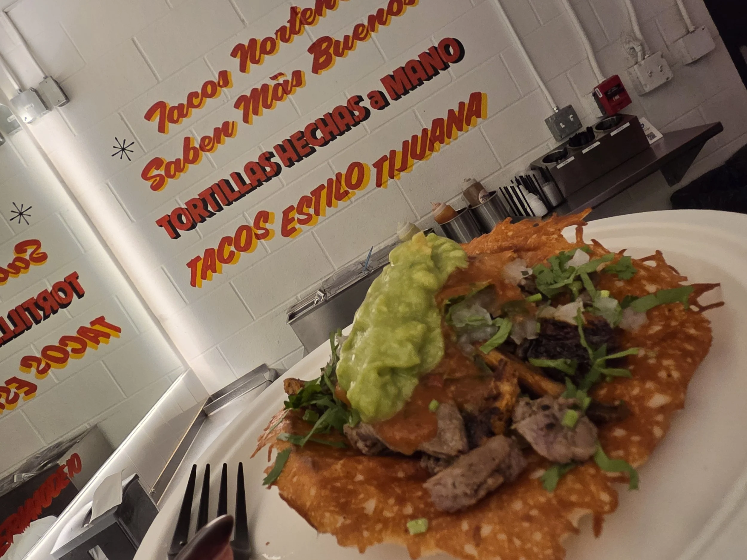 Close-up of a plate filled with Mexican food, including beef tacos with shredded beef, lettuce, and guacamole, set against a white brick wall with a red and yellow sign in Spanish.