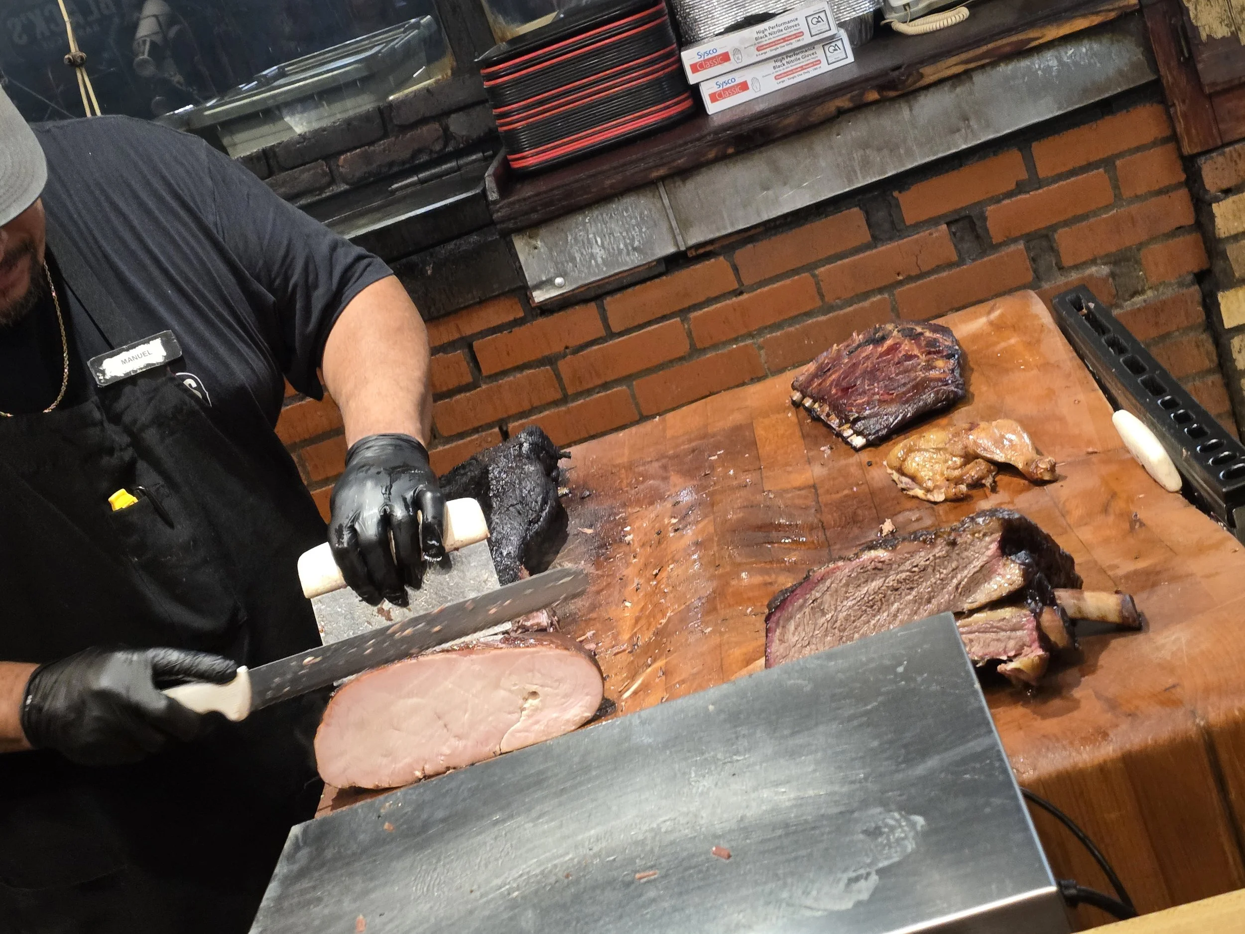 A bbq pitmaster wearing black gloves and  slicing cooked meat on a wooden cutting board in on a butcher board with brick walls.