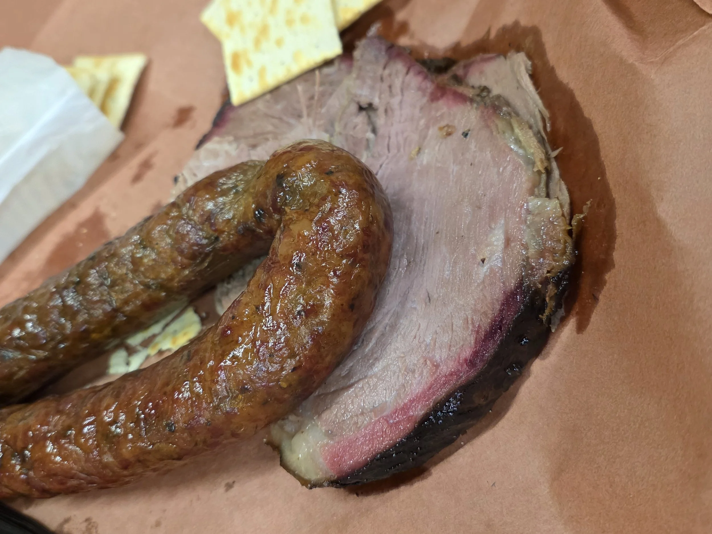 A close-up of a smoked piece of smoked shoulder clod with a sausage on top, served on butcher paper. There are some crackers in the background.