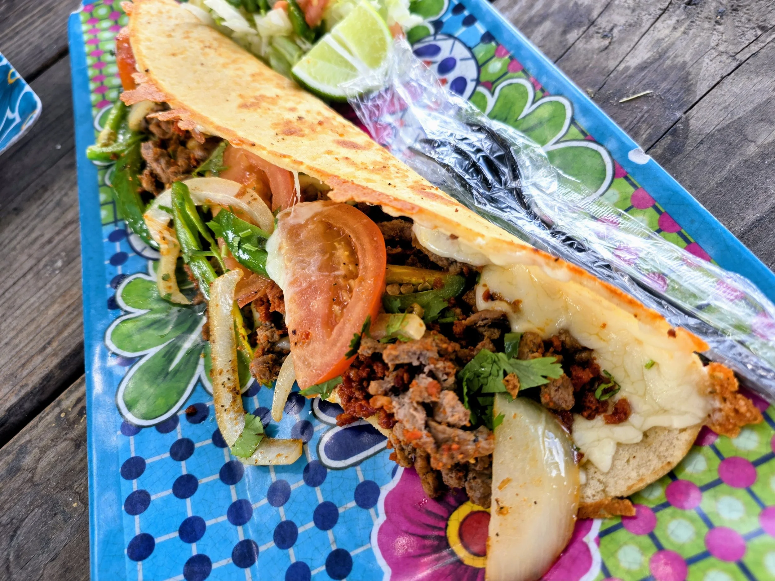 Close-up of a Mexican taco with ground beef, fresh vegetables, tomato, onion, cilantro, lime wedges, and cheese on a colorful paper plate on a wooden surface.