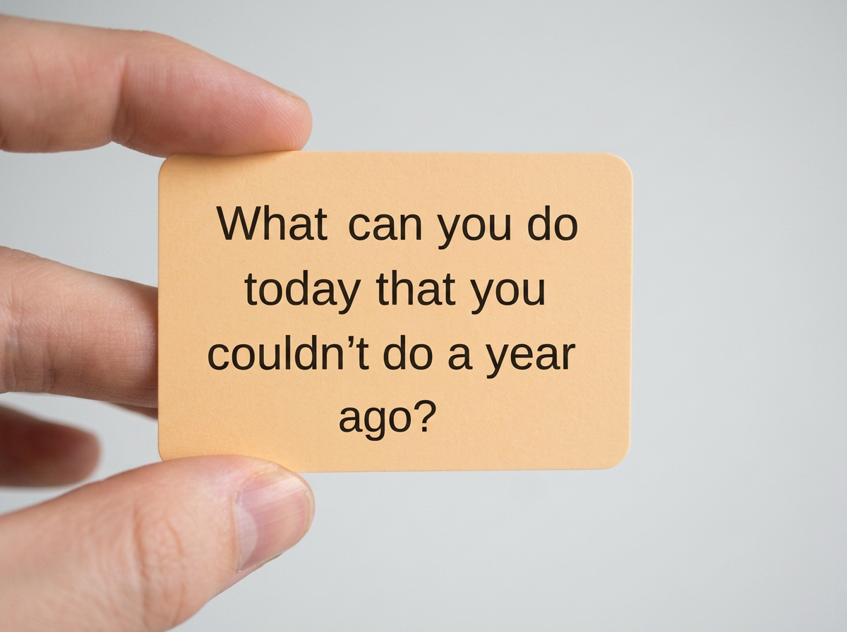 A hand holding a peach-colored card with the text, "What can you do today that you couldn’t do a year ago?" against a light background.