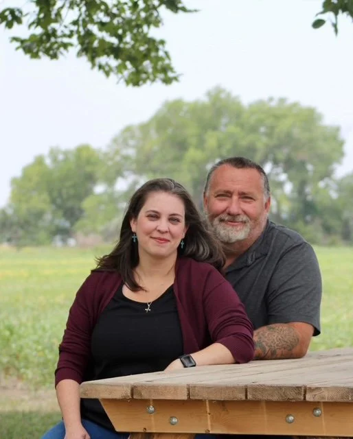 A woman and a man sitting outdoors at a wooden picnic table, smiling, with trees and greenery in the background.