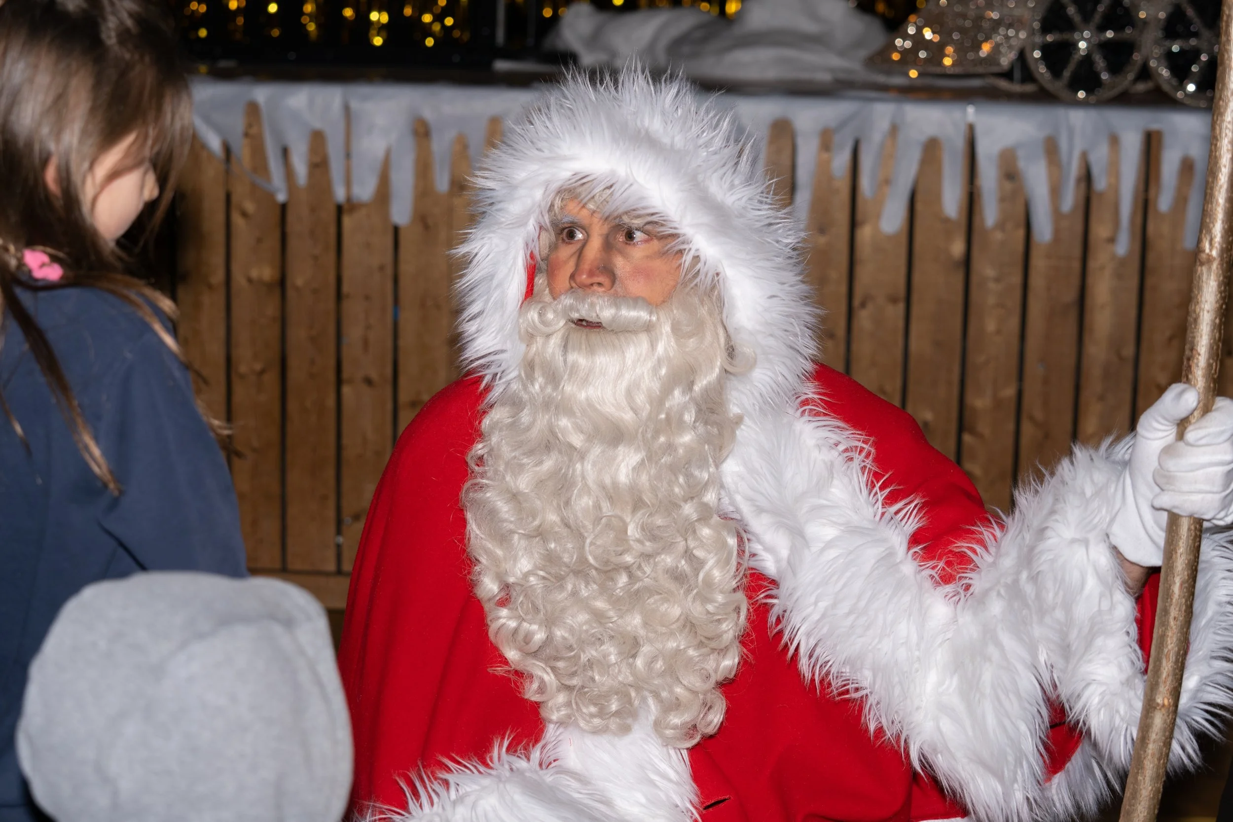 Santa Claus with a white beard and red suit sitting with a serious expression, talking to a young girl in a winter coat, with a wooden fence and Christmas decorations in the background.