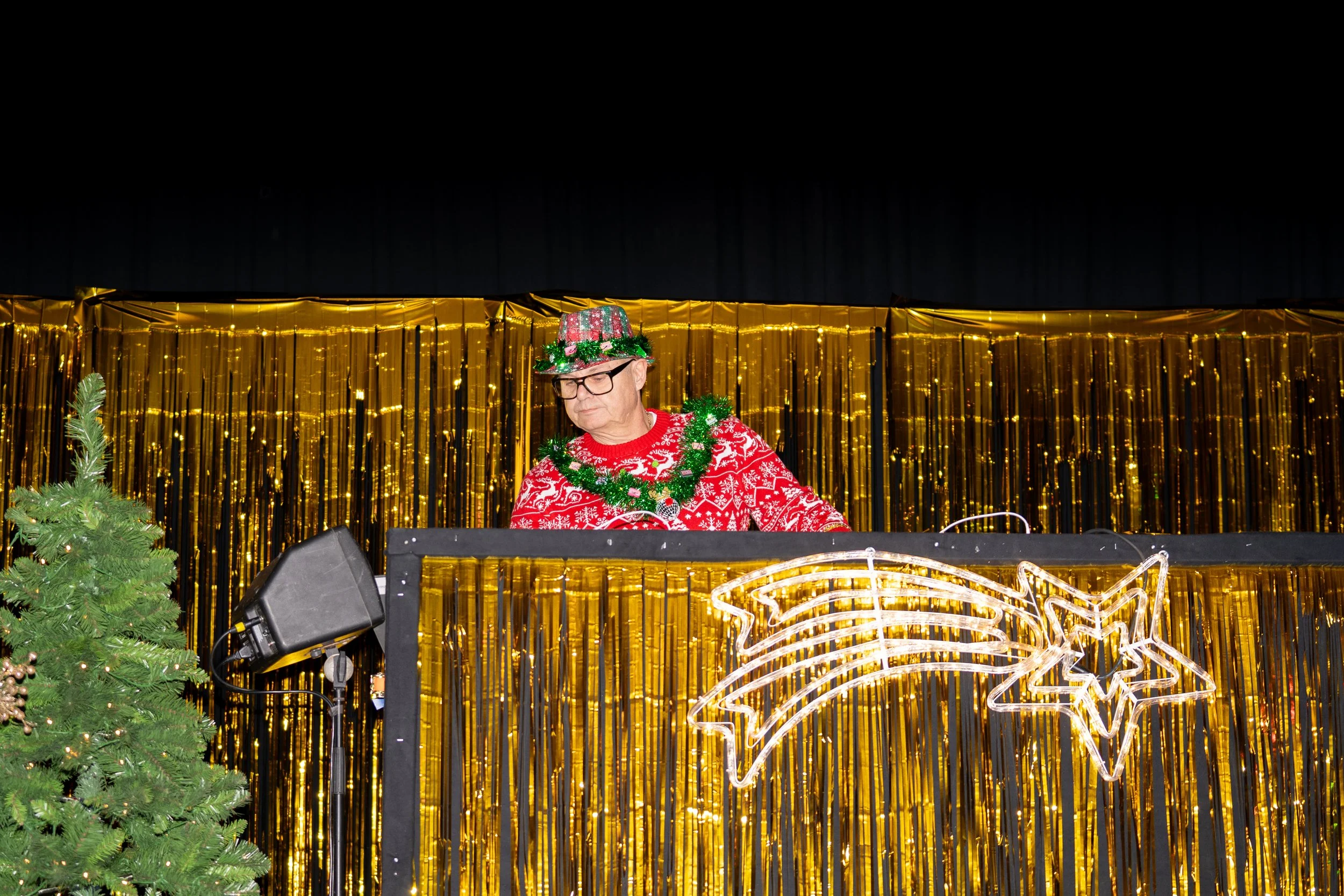 A man with glasses dressed in a red and white Christmas sweater wearing a festive hat decorated with holly leaves, standing at a DJ booth with Christmas lights and a gold backdrop.