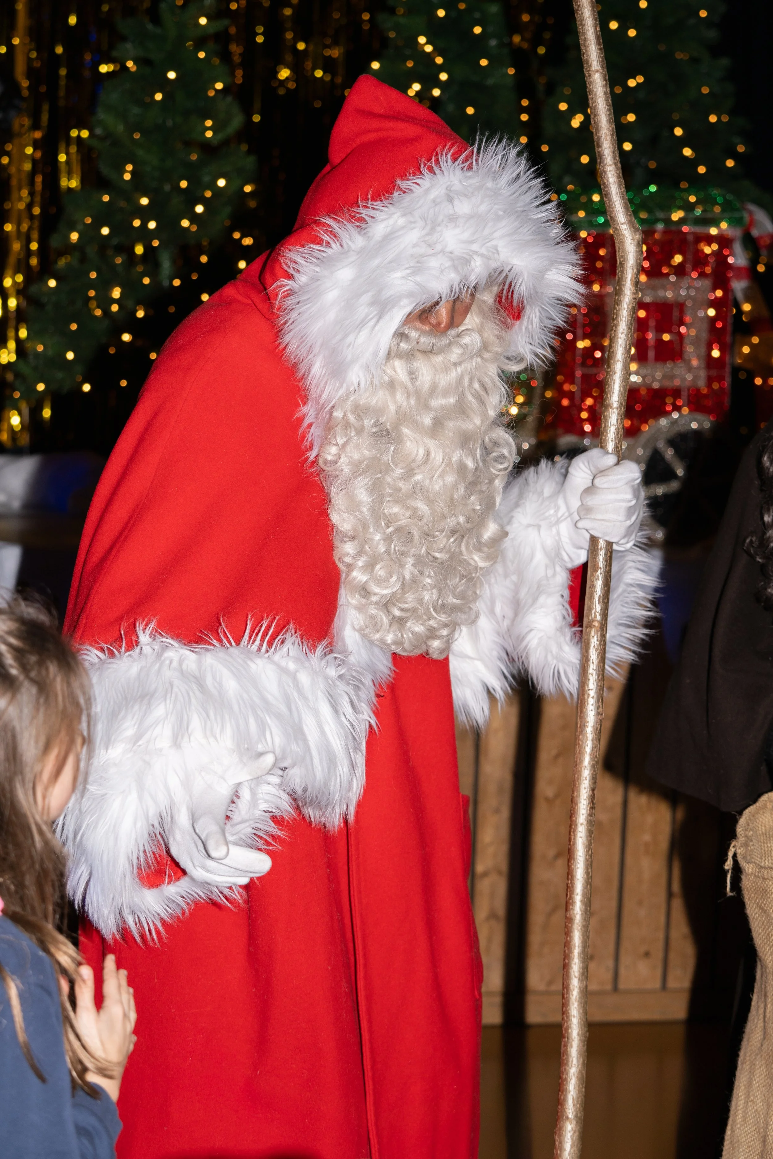 Santa Claus dressed in traditional red and white costume, holding a staff, with children nearby, in front of a decorated Christmas tree with lights.