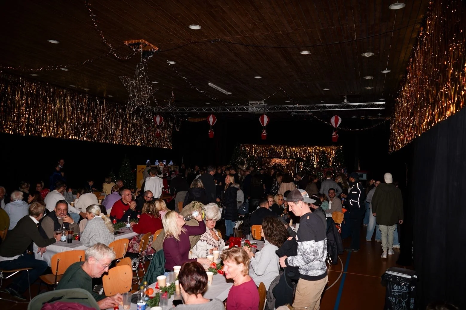 A crowded indoor Christmas celebration with decorated holiday lights, hanging paper lanterns, and festive decorations. People are gathered around tables, eating, drinking, and chatting, with a stage or performance area visible in the background.