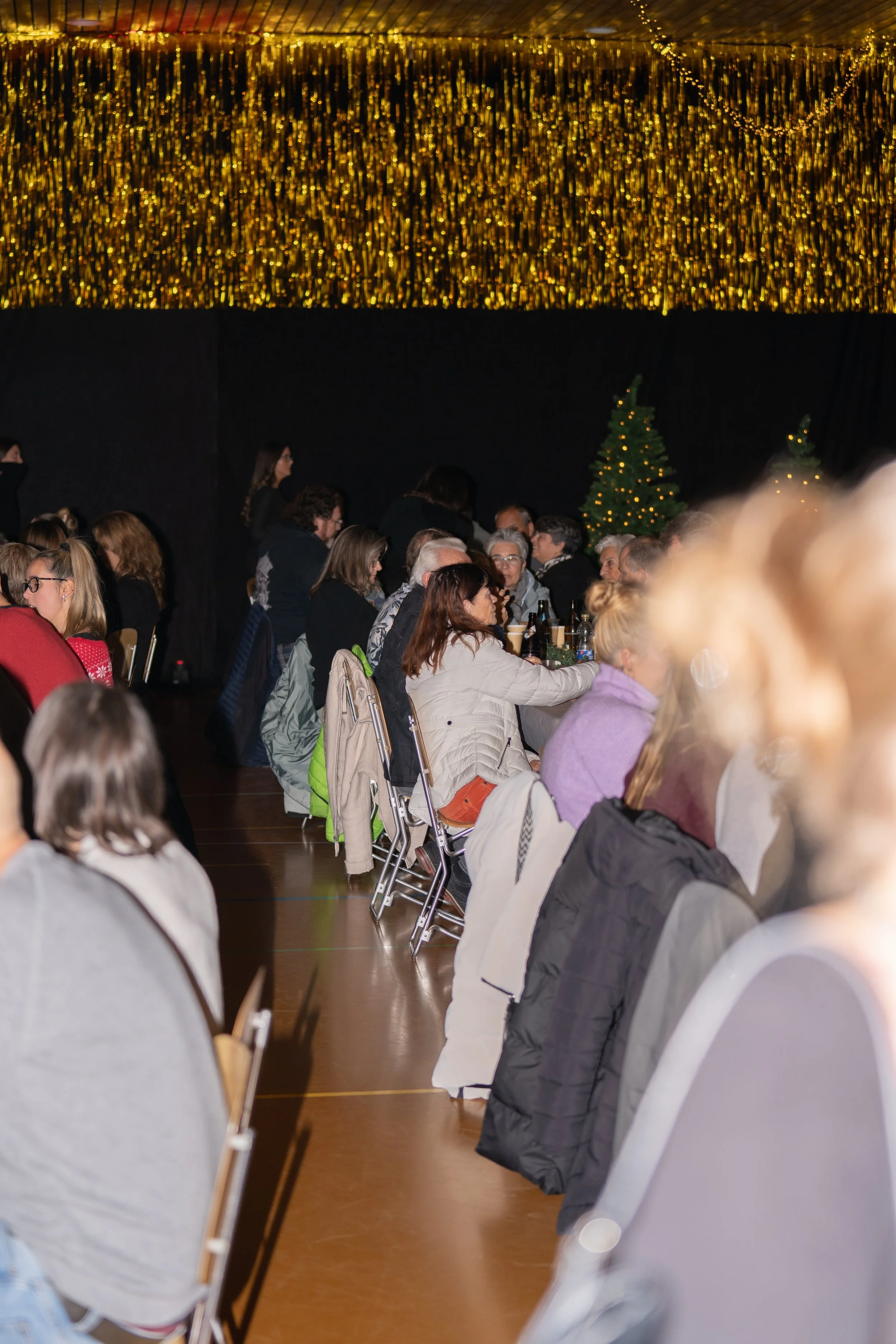 People seated at long tables in a decorated hall with Christmas trees and golden streamers hanging from the ceiling, likely at a holiday event or party.