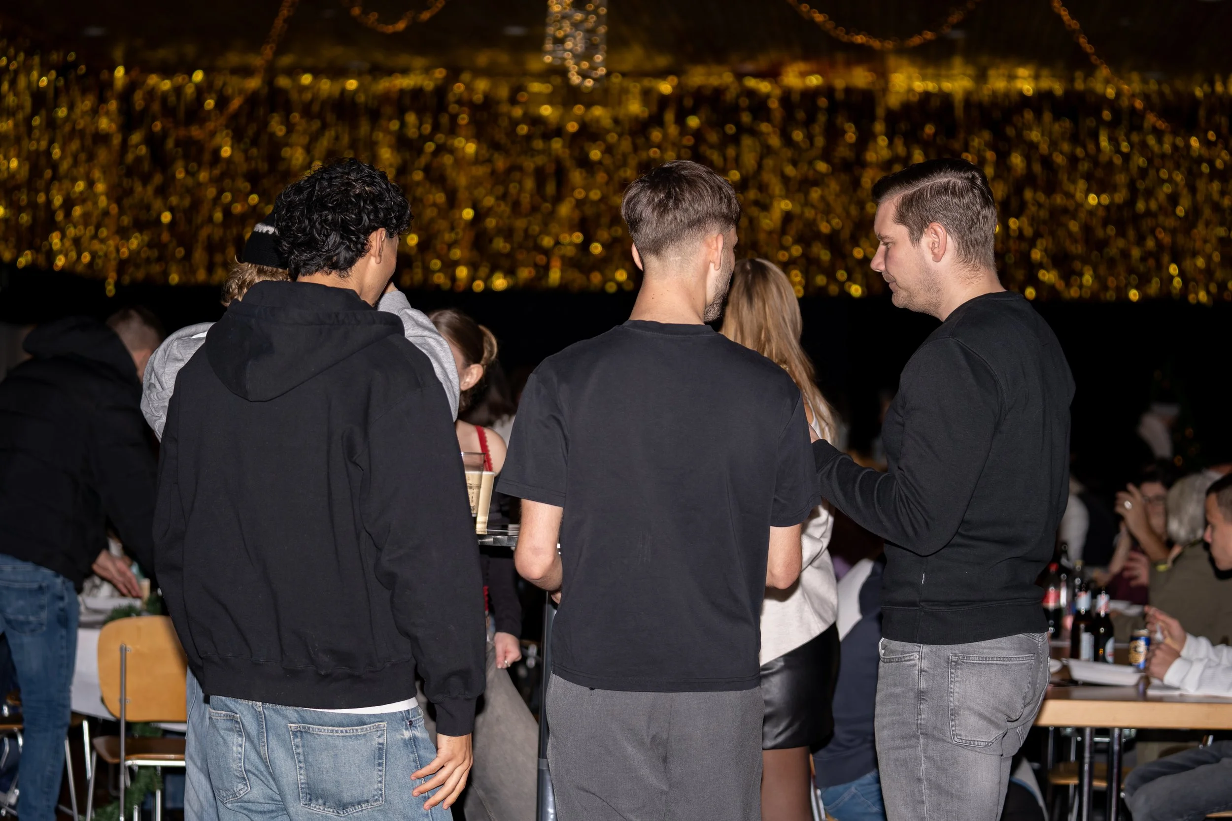 Group of people at a social gathering, talking and exchanging drinks, with festive lights in the background.