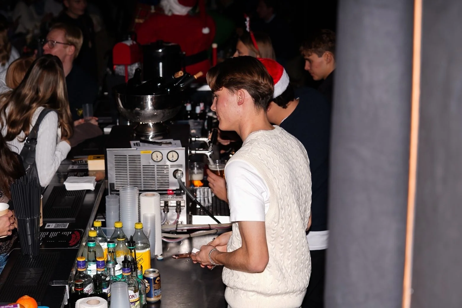 People gathered at a bar or party area with some wearing Christmas accessories like Santa hats.