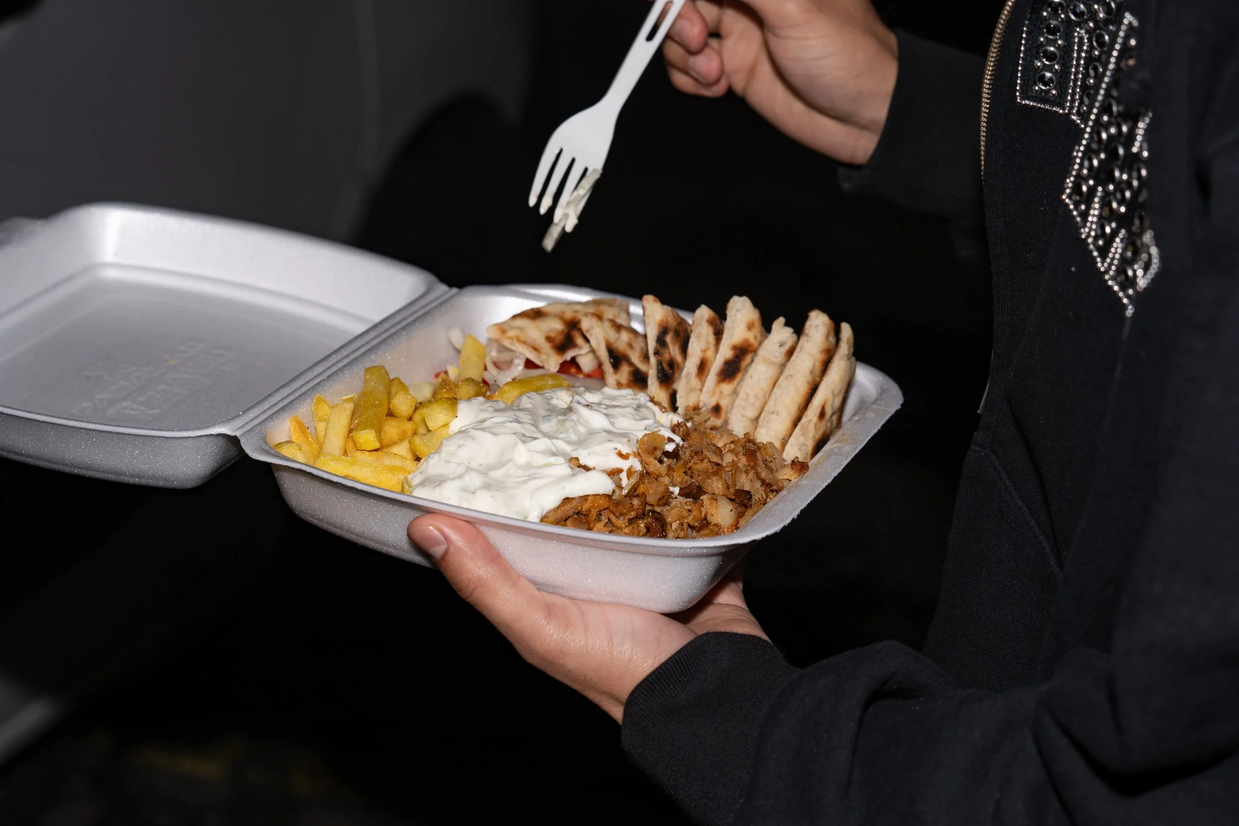 Person holding a styrofoam takeout container with food, including fries, grilled chicken, pita bread, and toppings, while using a plastic fork in their other hand.