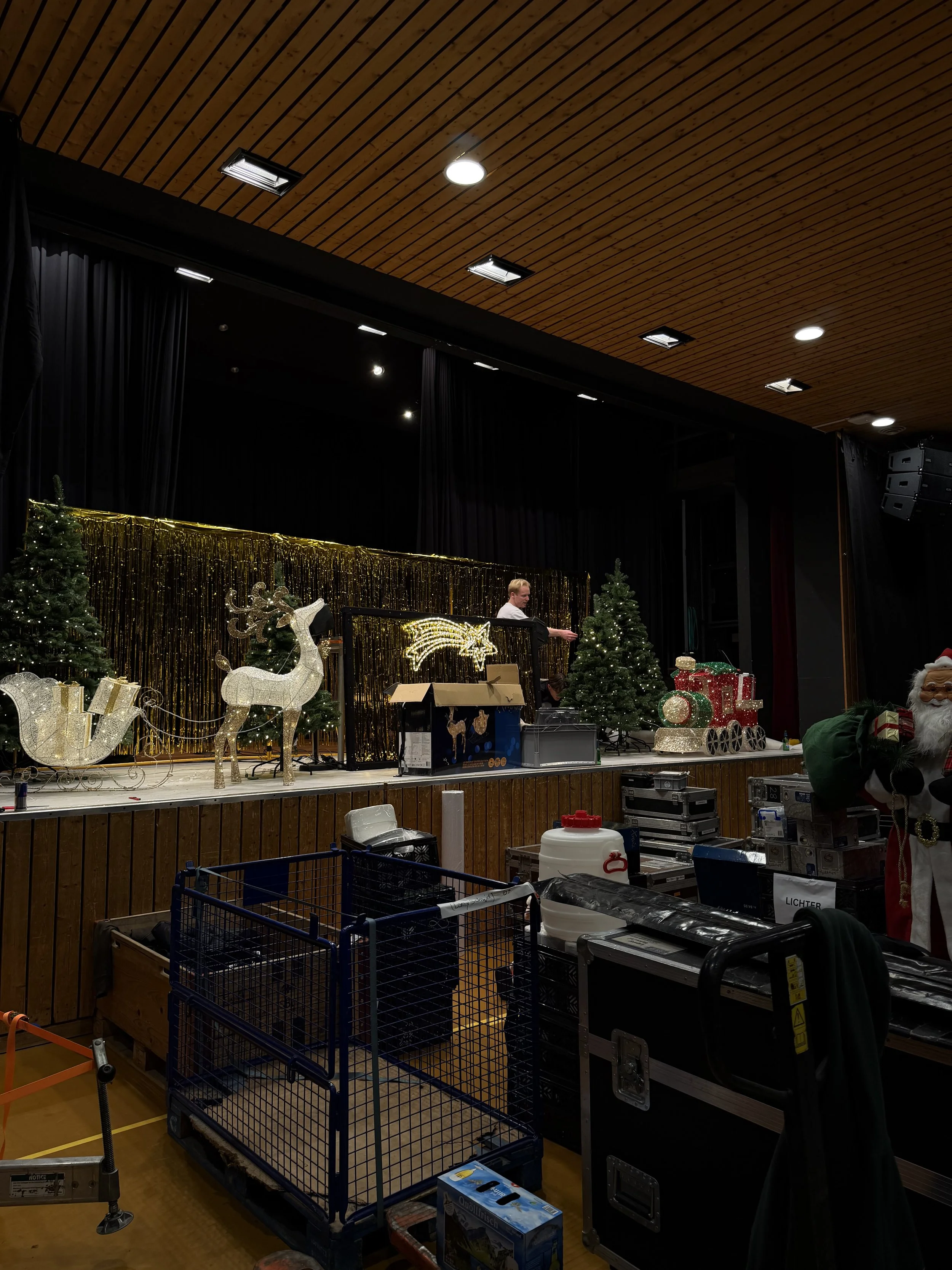 Stage with Christmas decorations, including Christmas trees, reindeer, and gift boxes, in a room with a wooden ceiling and black curtains.