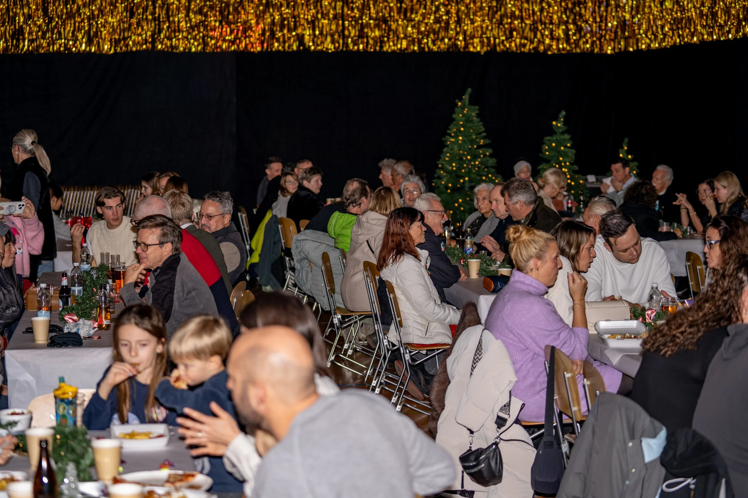 People gathered at decorated tables for a holiday celebration with Christmas trees in the background.