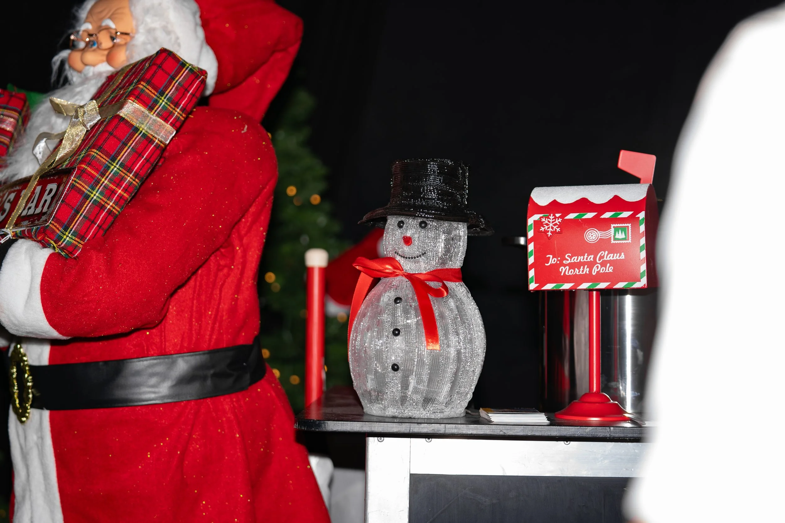 Decorative snowman figure with a black top hat and red ribbon, next to a red mailbox labeled 'Jo: Santa Claus North Pole,' and part of a figure in a red coat with a plaid gift box on its shoulder.
