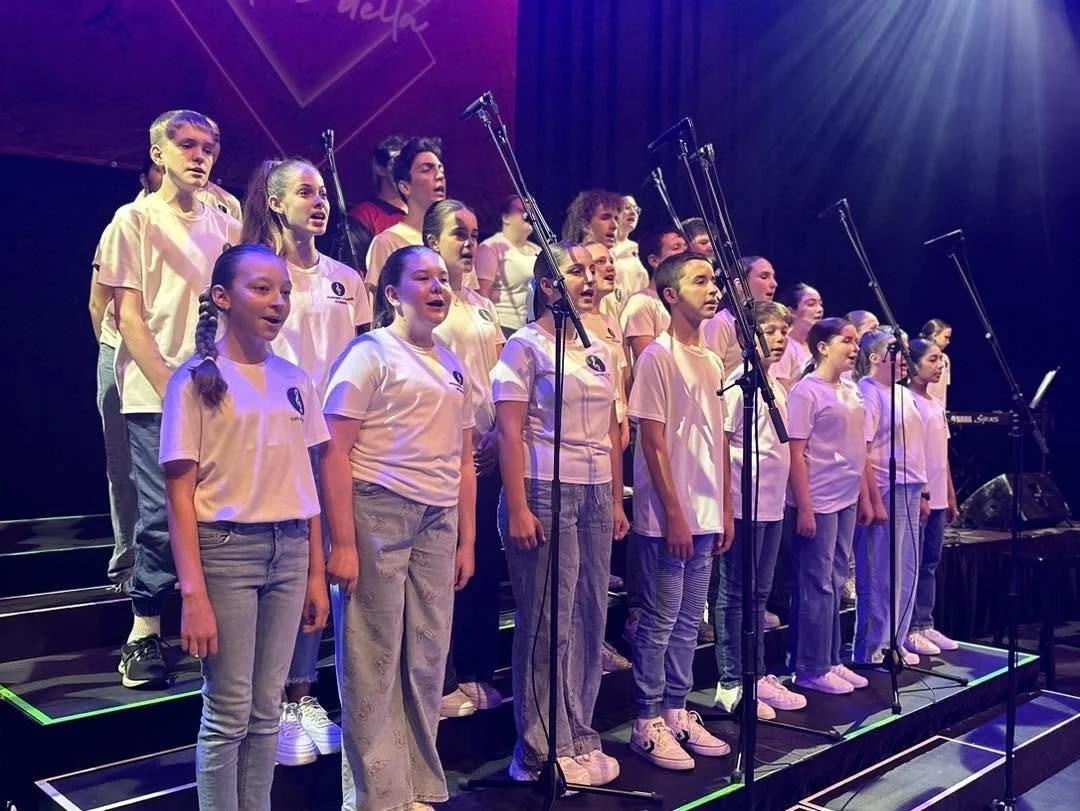 Group of children singing on stage under colorful lights, arranged in rows with microphones in front.