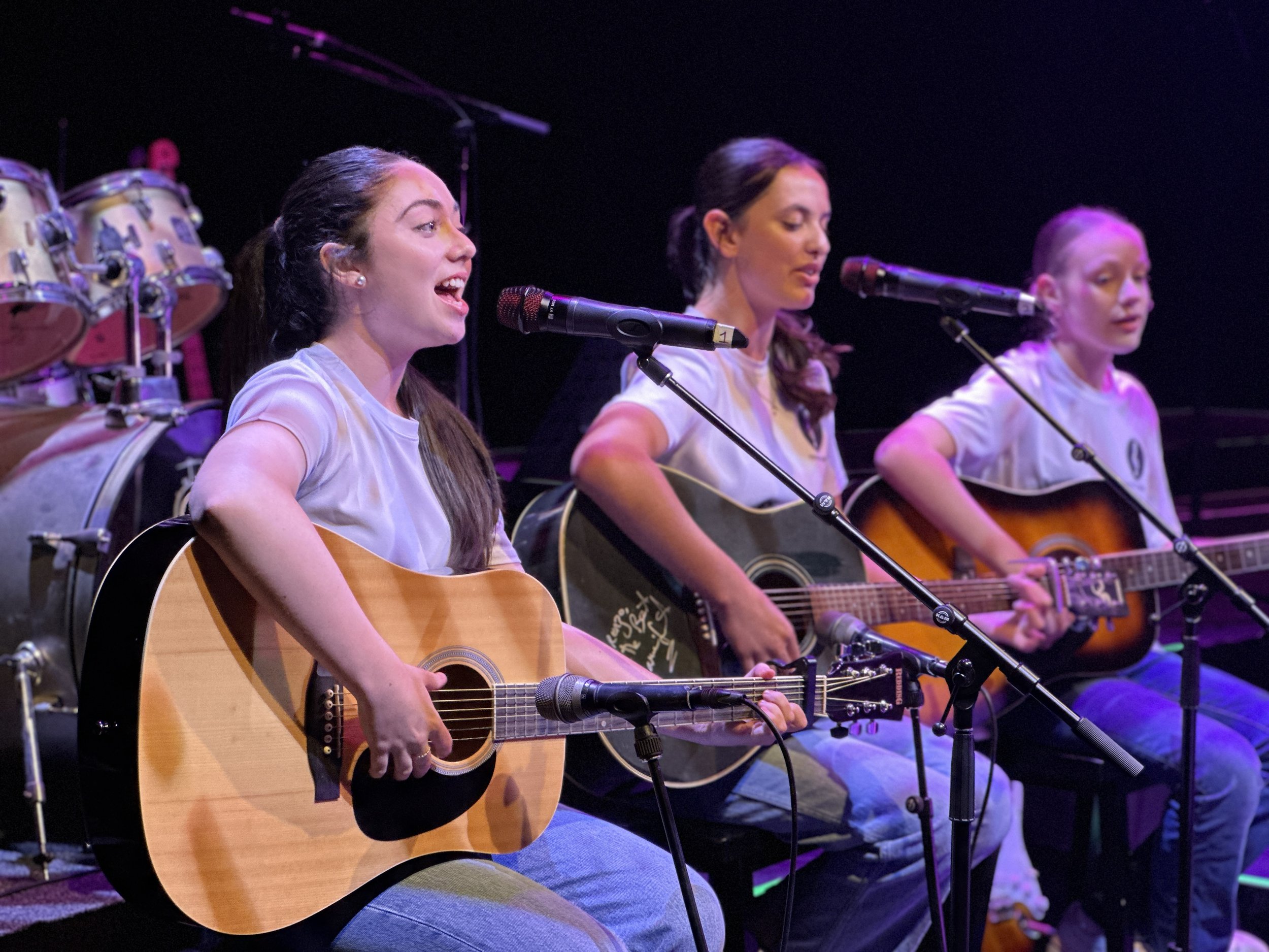 Three women sitting on stage playing acoustic guitars and singing into microphones, with drums in the background, illuminated by purple stage lighting.