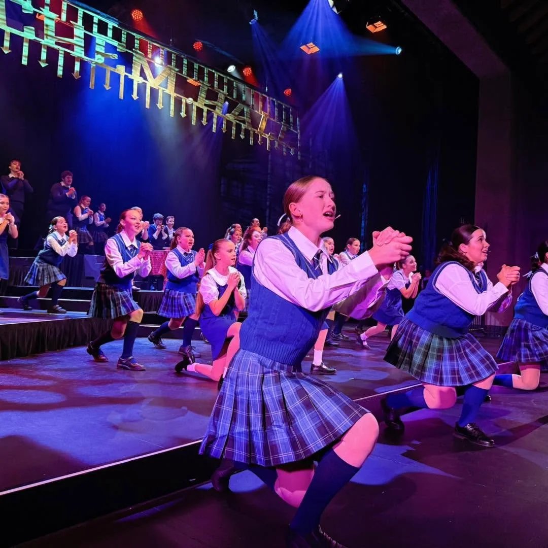 A group of young girls in school uniforms performing on stage during a theatrical production, with some kneeling and others standing, under colorful stage lighting.