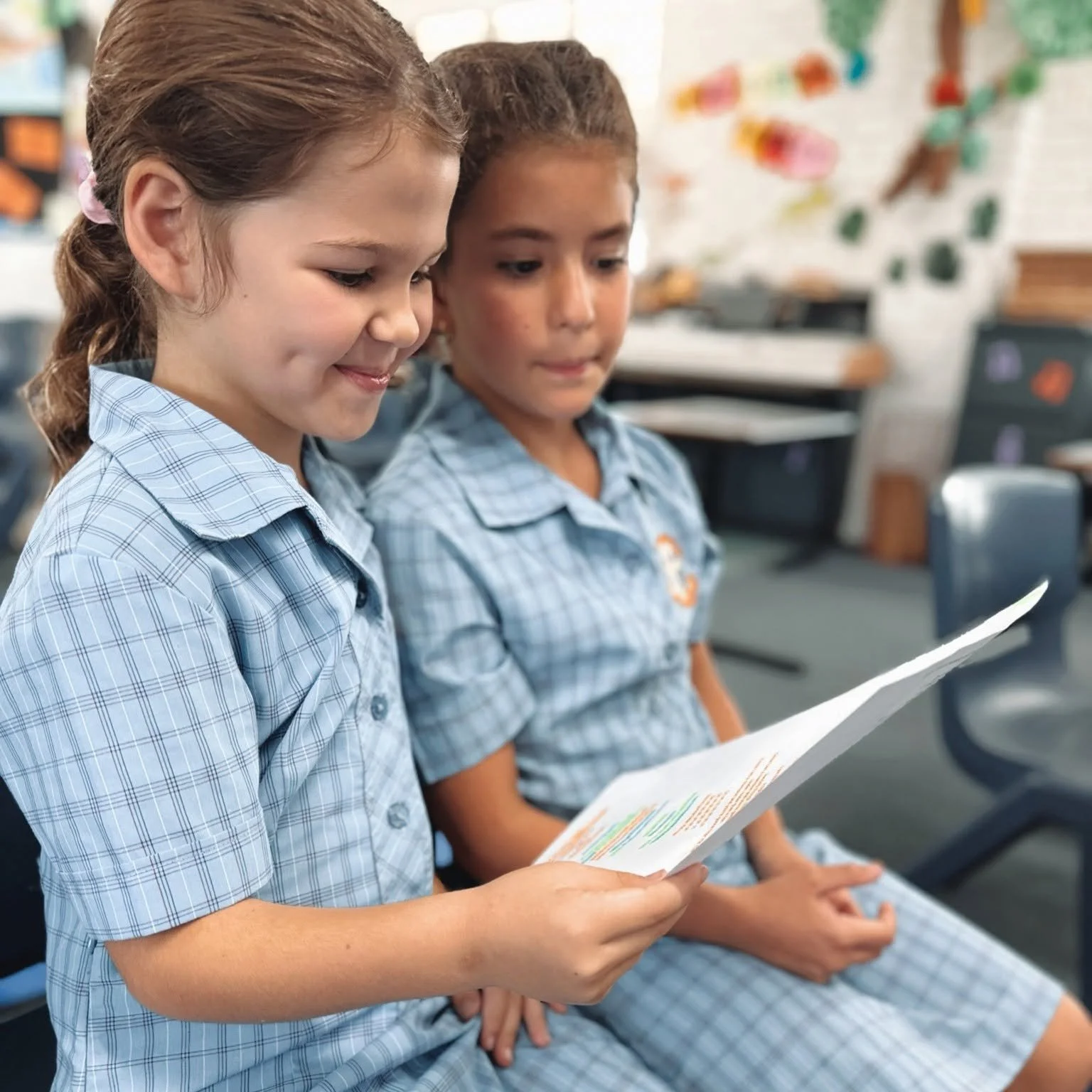 Two young girls in school uniforms sitting in a classroom, reading a paper together, with classroom decorations in the background.