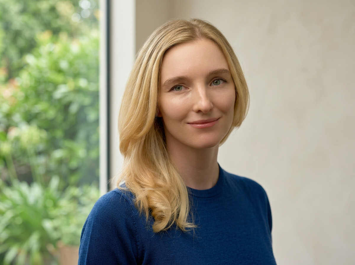 A young woman with long blonde hair smiling softly, wearing a blue top, standing indoors near a window with a green outdoor scene visible in the background.