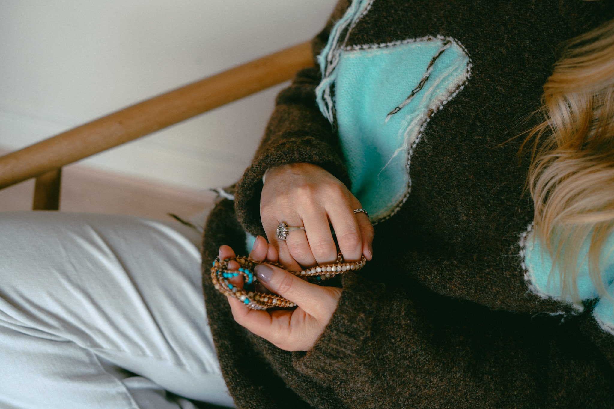 Close-up of a person holding beaded jewelry, wearing a turquoise face mask, a dark fuzzy sweater, and white pants, with long blonde hair in the background.