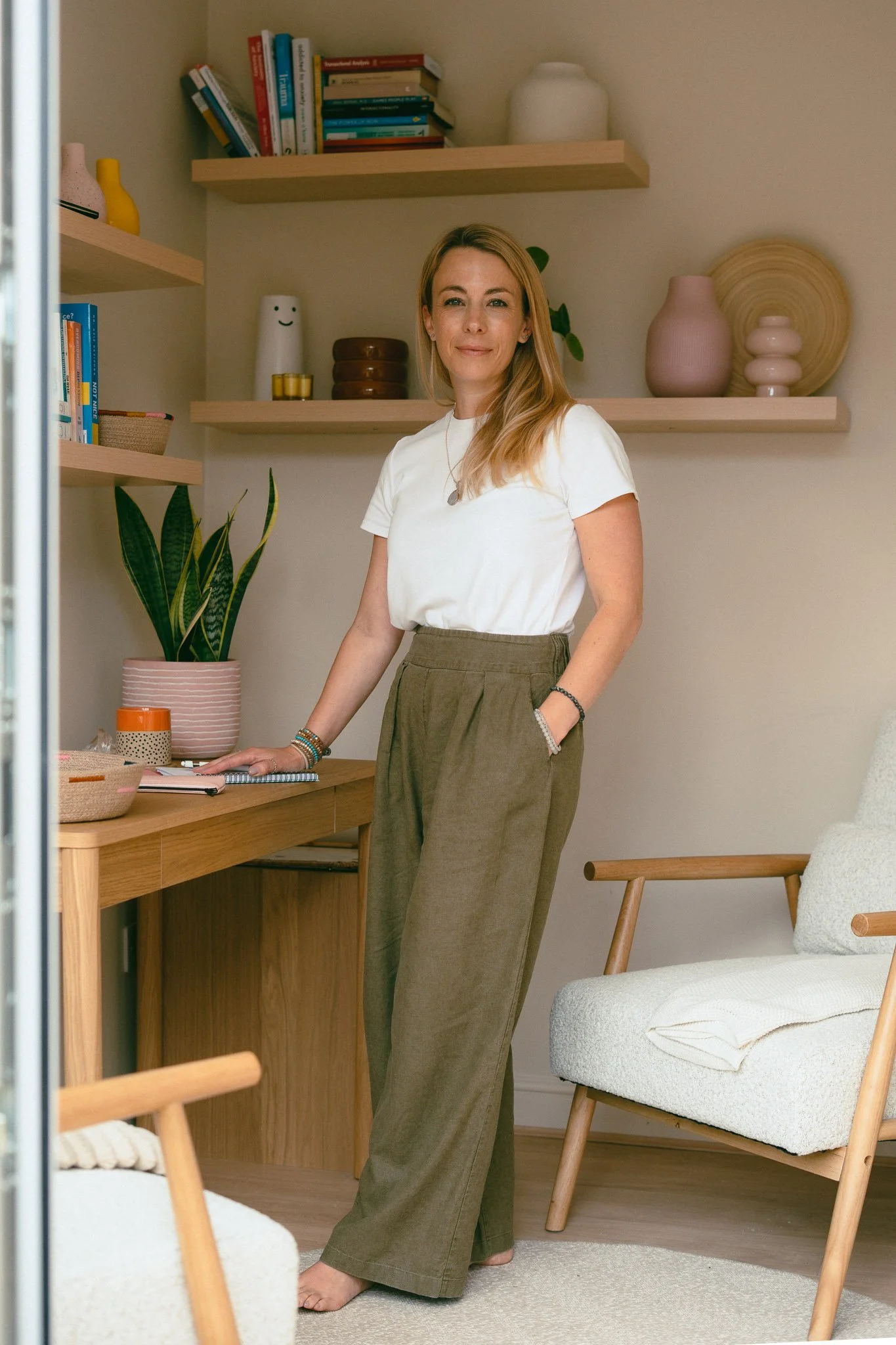A woman with blonde hair wearing a white T-shirt and khaki pants, standing in a cozy, modern living space with wooden furniture, shelves decorated with vases, books, plants, and art, smiling at the camera.
