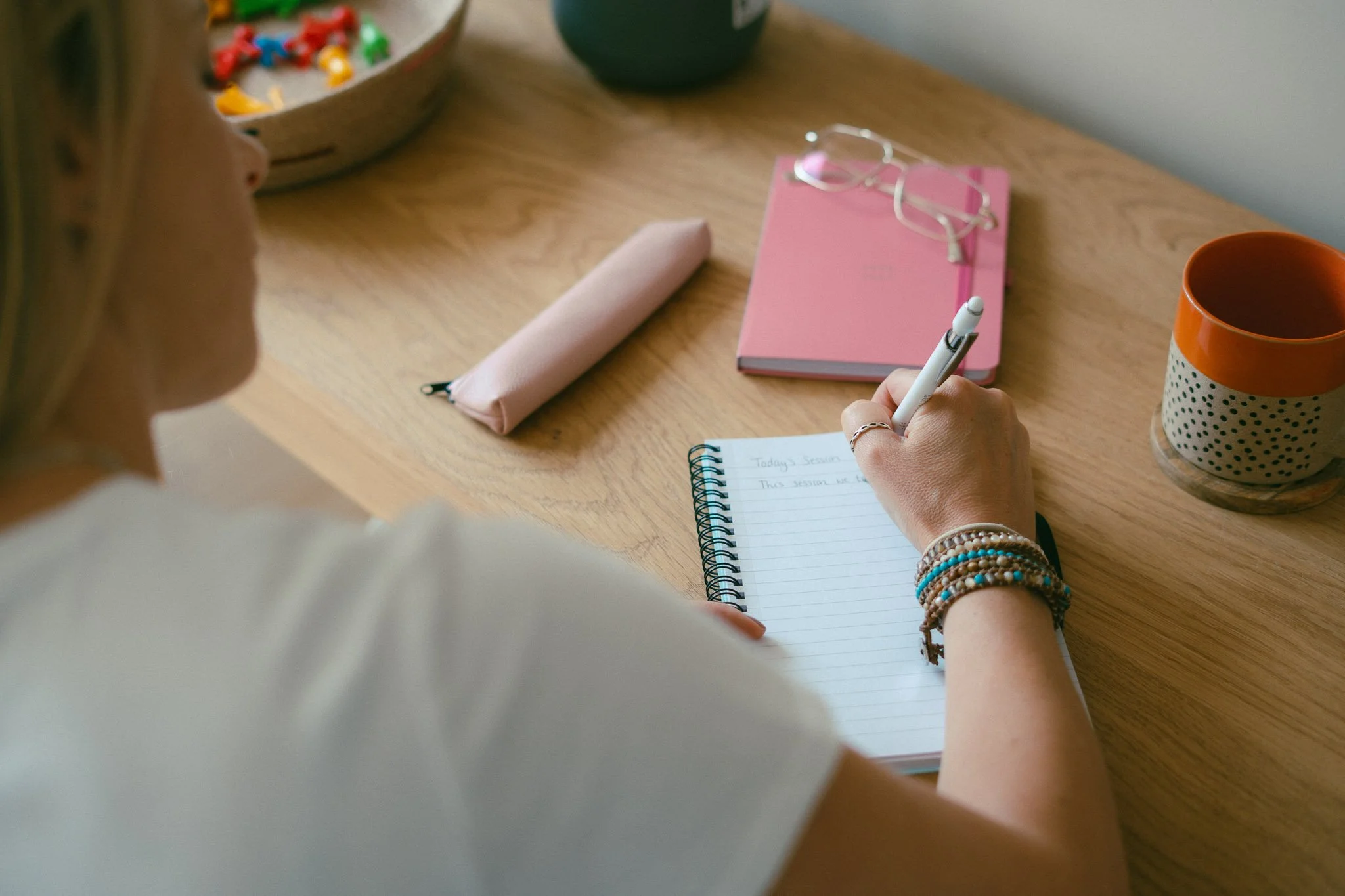 Person writing in a spiral notebook at a wooden desk with a pink notebook, glasses, a striped mug, and a pouch.