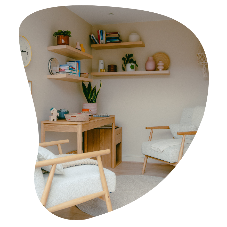 A cozy living room corner with light-colored walls and wooden furniture, including a white armchair and a small wooden desk with decorative items, potted plants, and floating shelves holding books and pottery.